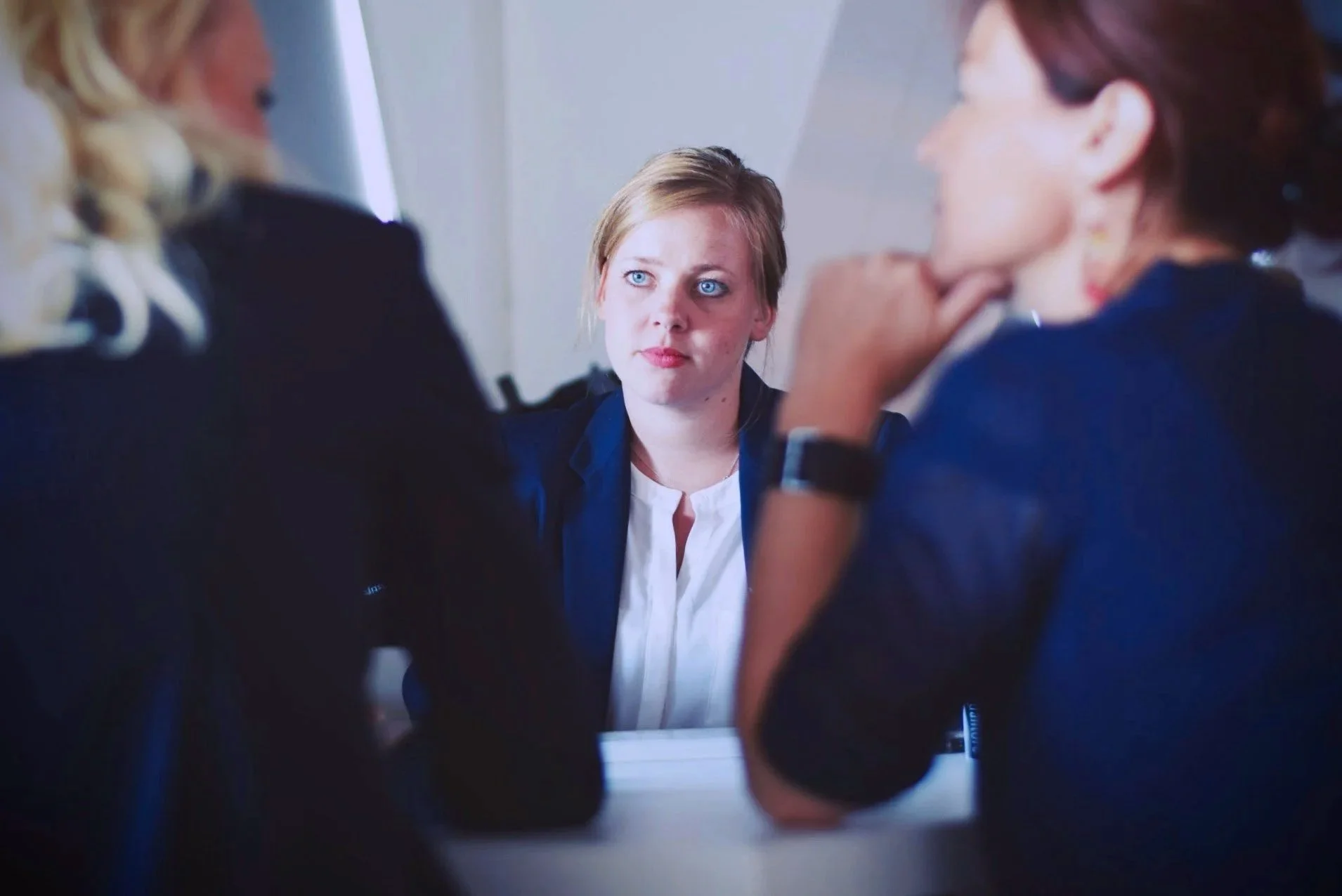 Three women are engaged in a serious discussion, one with blonde hair and two with red hair, in a professional setting.