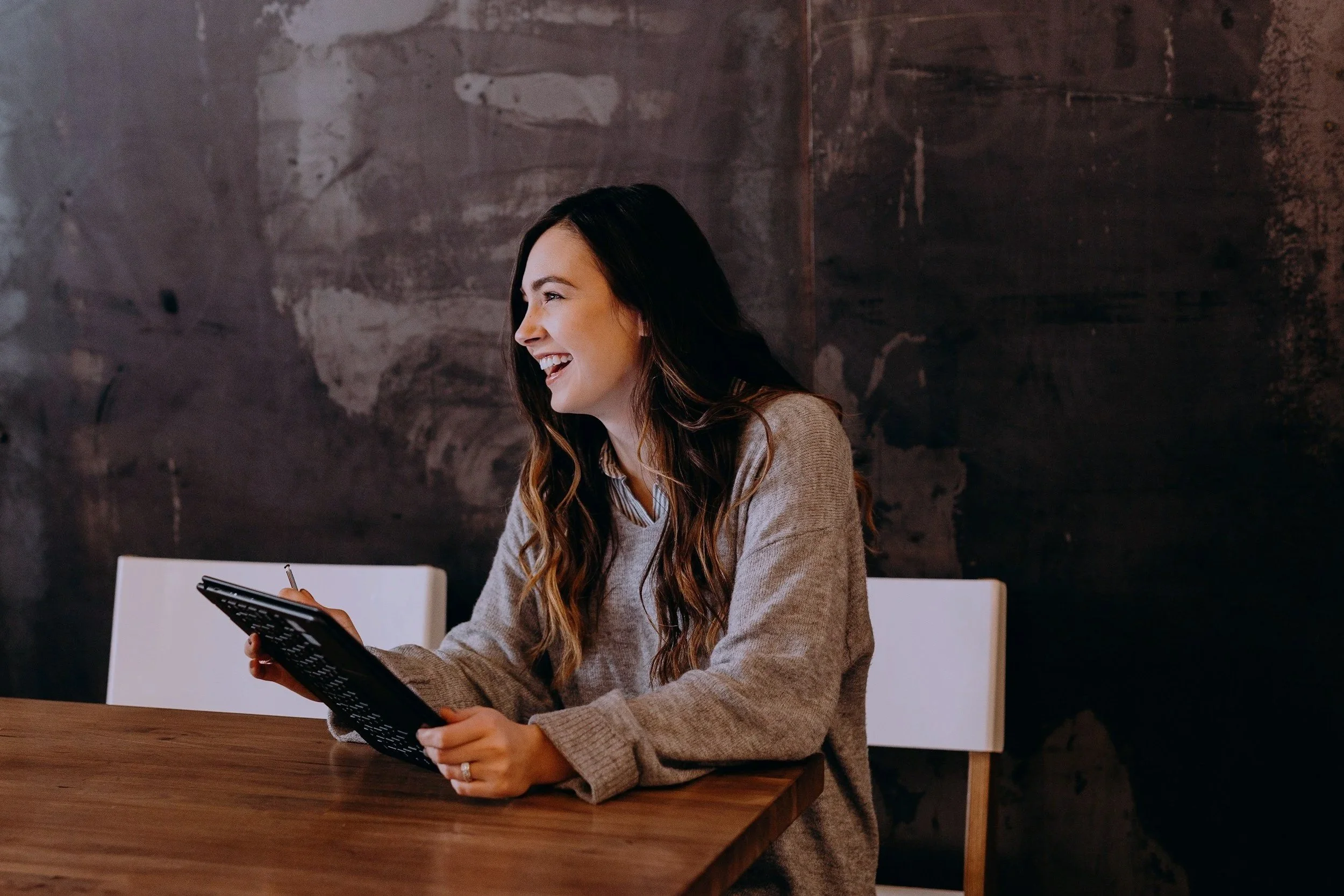 A woman with long brown hair wearing a beige sweater, sitting at a wooden table, smiling and looking to her left while holding a black tablet device.