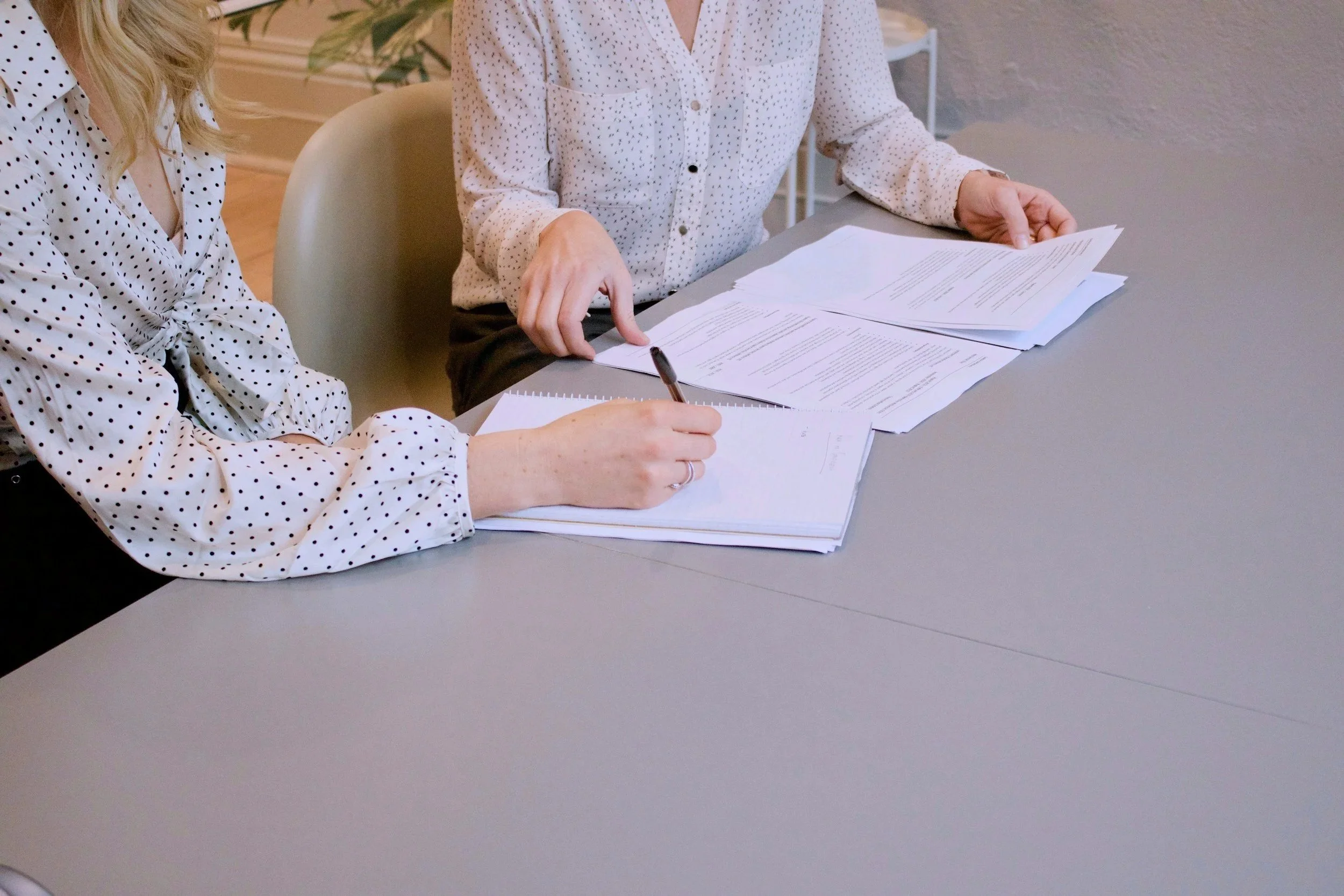 Two women sitting at a table reviewing documents and taking notes, both dressed in white blouses with black polka dots.