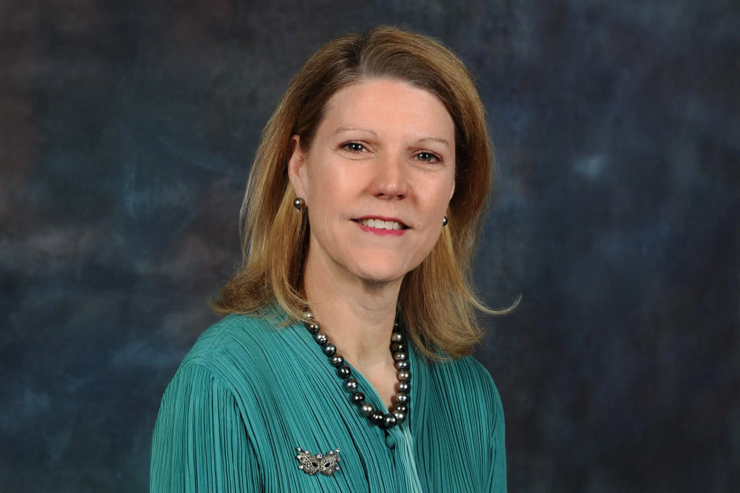 A middle-aged woman with light skin and shoulder-length reddish-blonde hair, smiling slightly, wearing a teal textured top, a black pearl necklace, earrings, and a decorative brooch, against a dark, cloudy background.