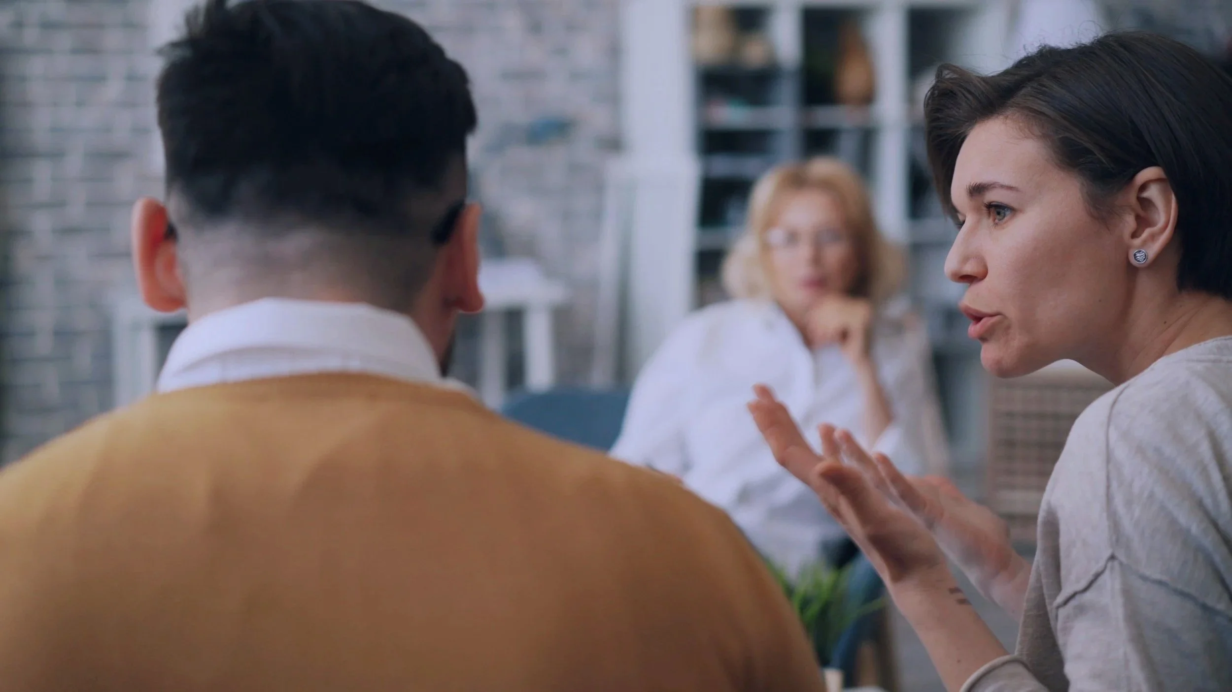 A woman with dark hair and earrings is speaking to a man with short dark hair who is wearing a yellow sweater. In the background, a blonde woman with glasses is sitting and listening.
