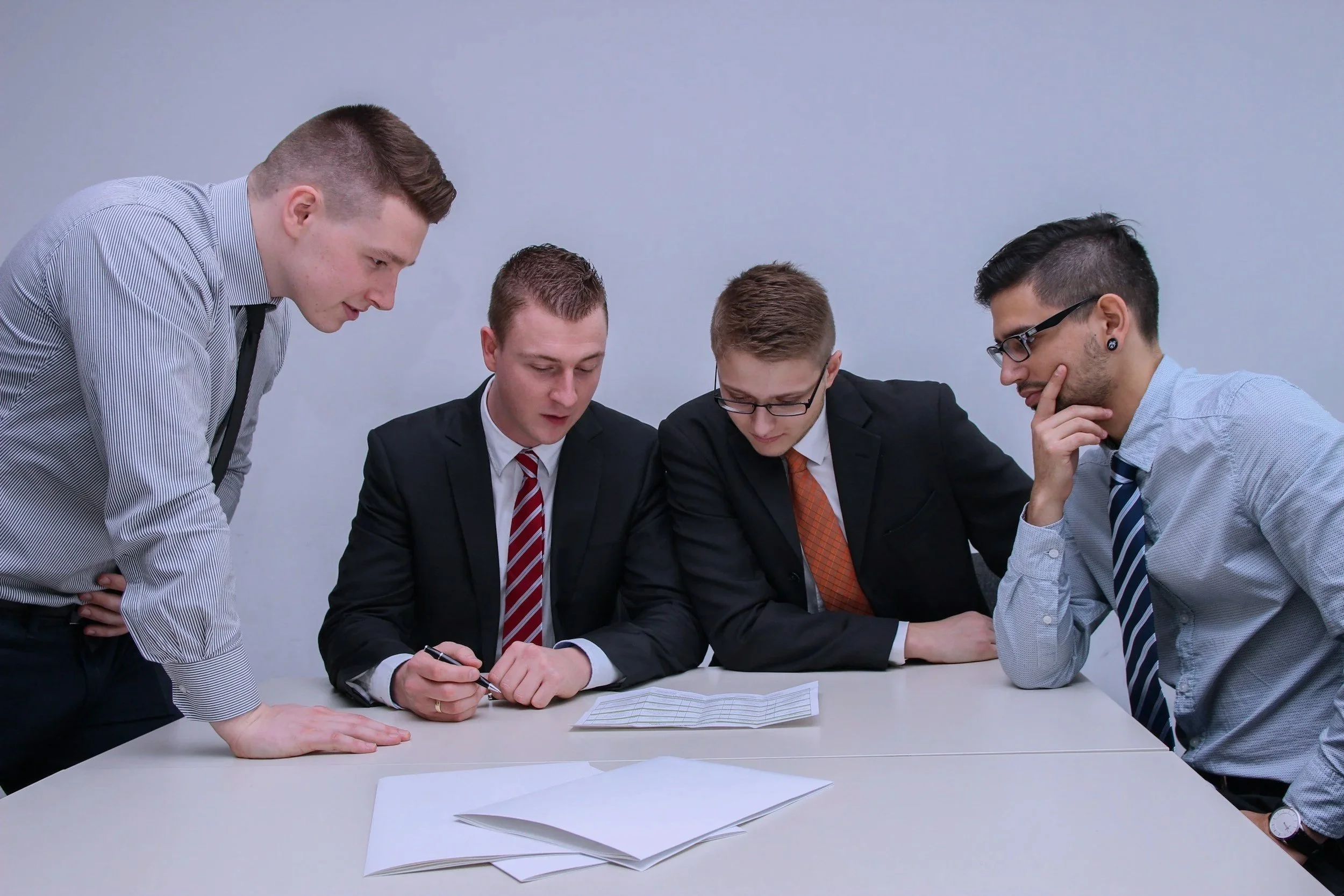 Four men in business attire gathered around a table reviewing documents, with one man signing papers while the others observe closely.