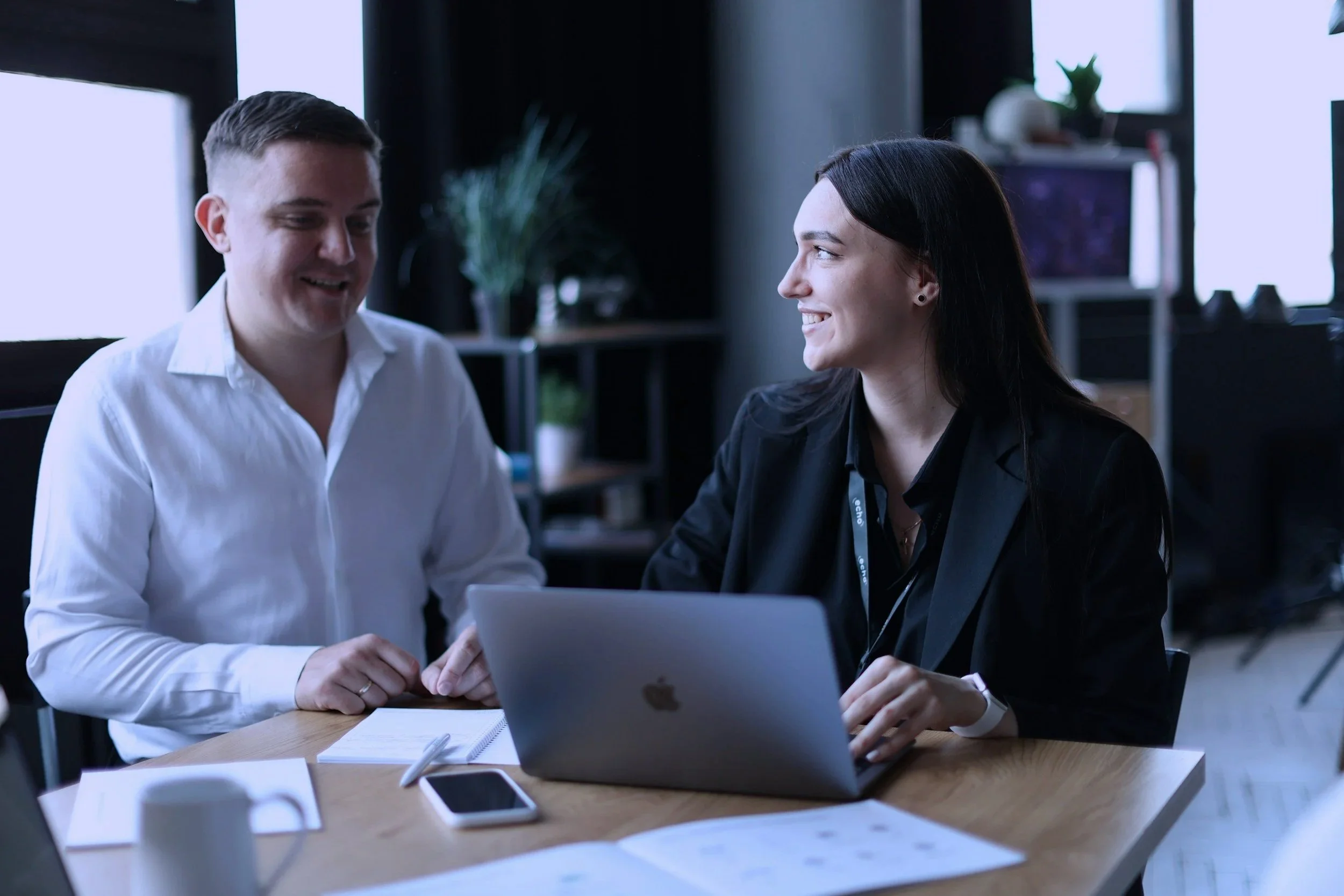 Two business professionals, a man and a woman, are sitting at a desk in an office, smiling and working with a laptop, with notebooks, a smartphone, and papers on the desk.