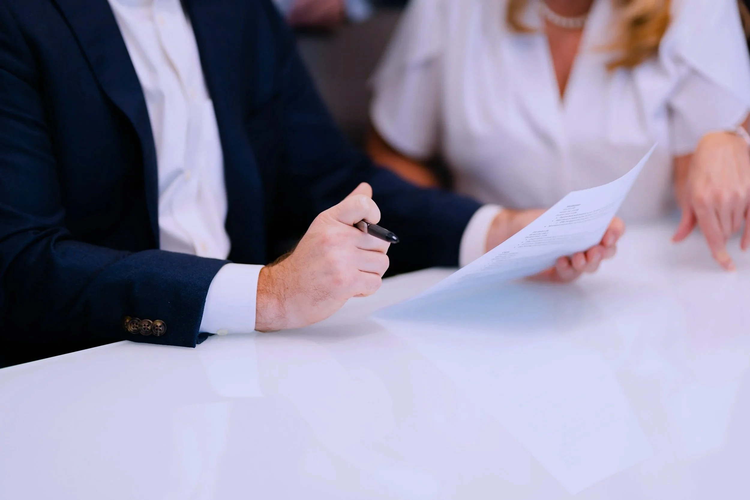 Close-up of a person wearing a dark blazer and white shirt, holding a pen and a document, sitting at a white table with another person in a white shirt in the background.