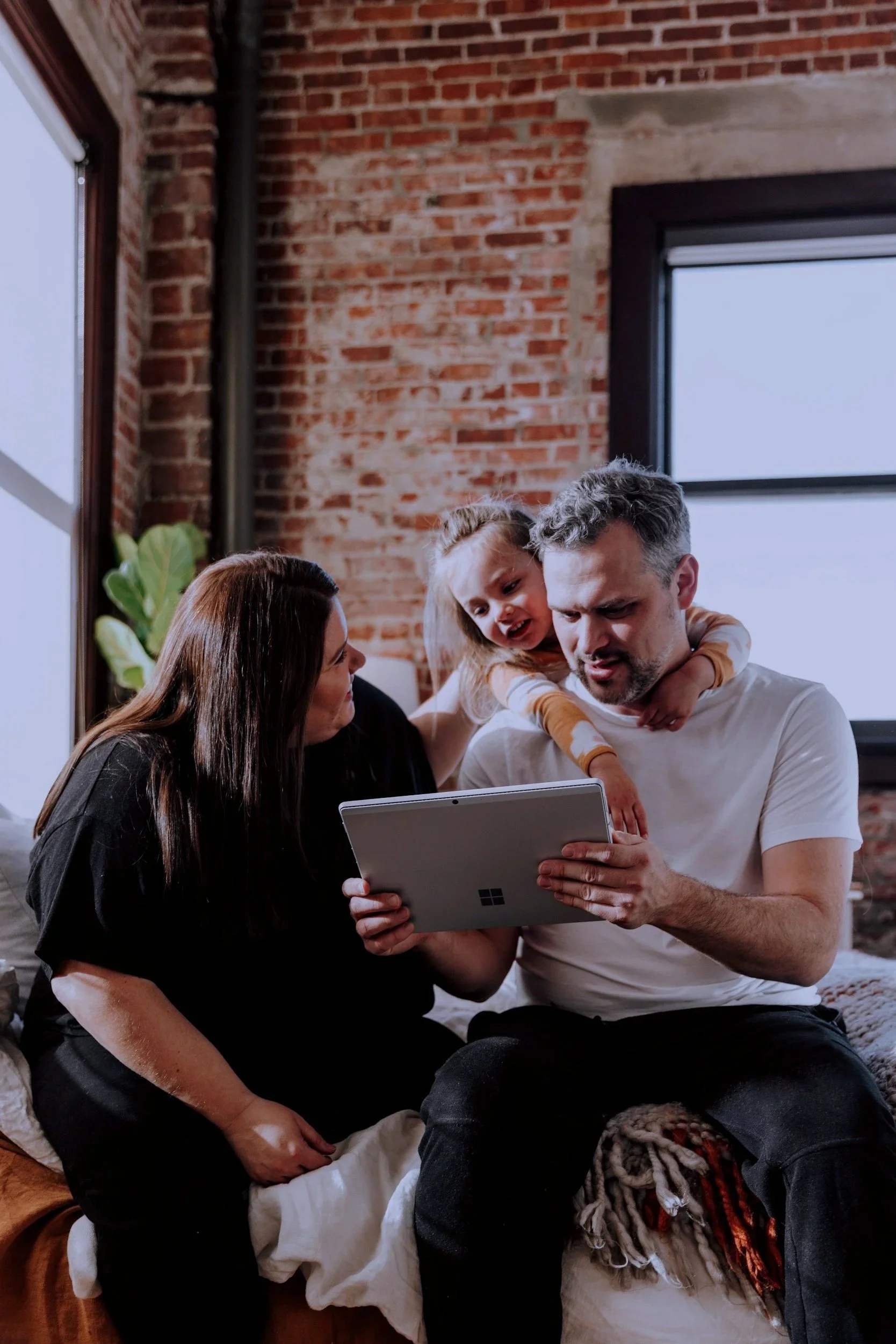 A family of three, consisting of a man, woman, and a young girl, sitting on a bed with a dog. The man is holding a tablet, the woman is sitting beside them, and the girl is on the man's shoulders, looking at the tablet. The room has a brick wall, large window, and indoor plants.