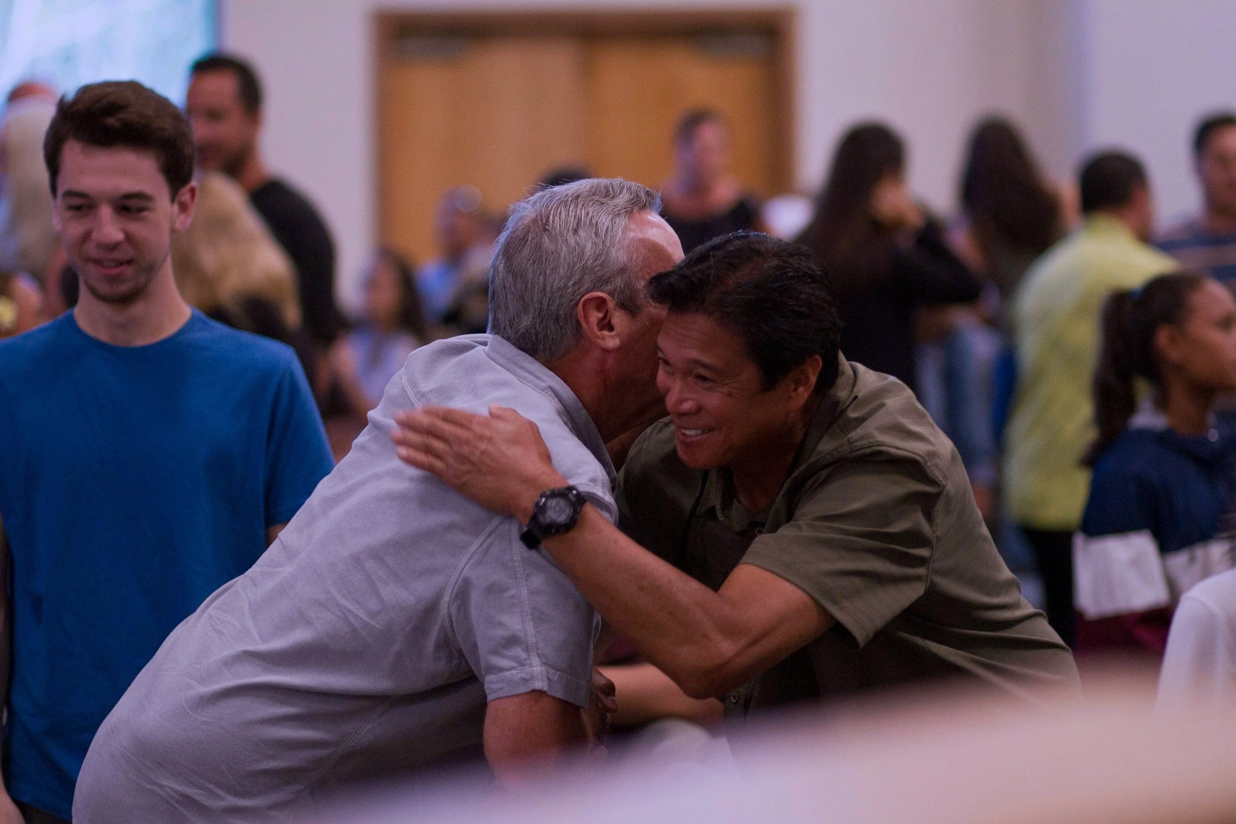 Two men hugging, smiling, in a crowded indoor setting with several people in the background.