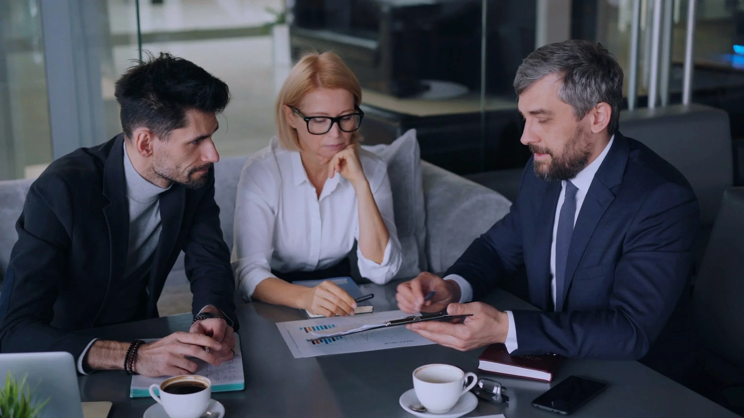 Three business professionals, two men and one woman, engaged in a serious discussion around a table in a modern office. They are reviewing documents, with two cups of coffee and a smartphone on the table.