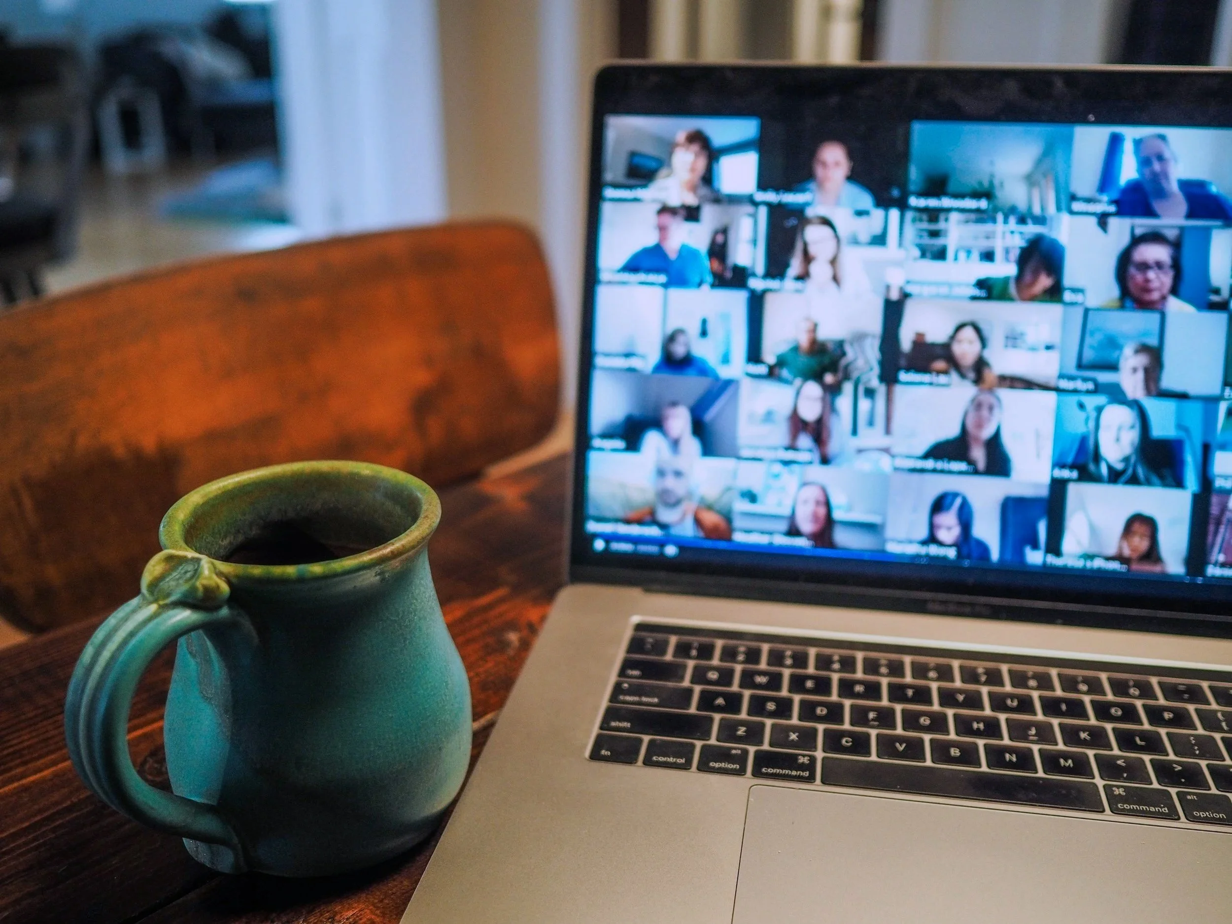 A computer screen showing a video conference with multiple participants, a coffee mug, and a ceramic teapot on a wooden table.