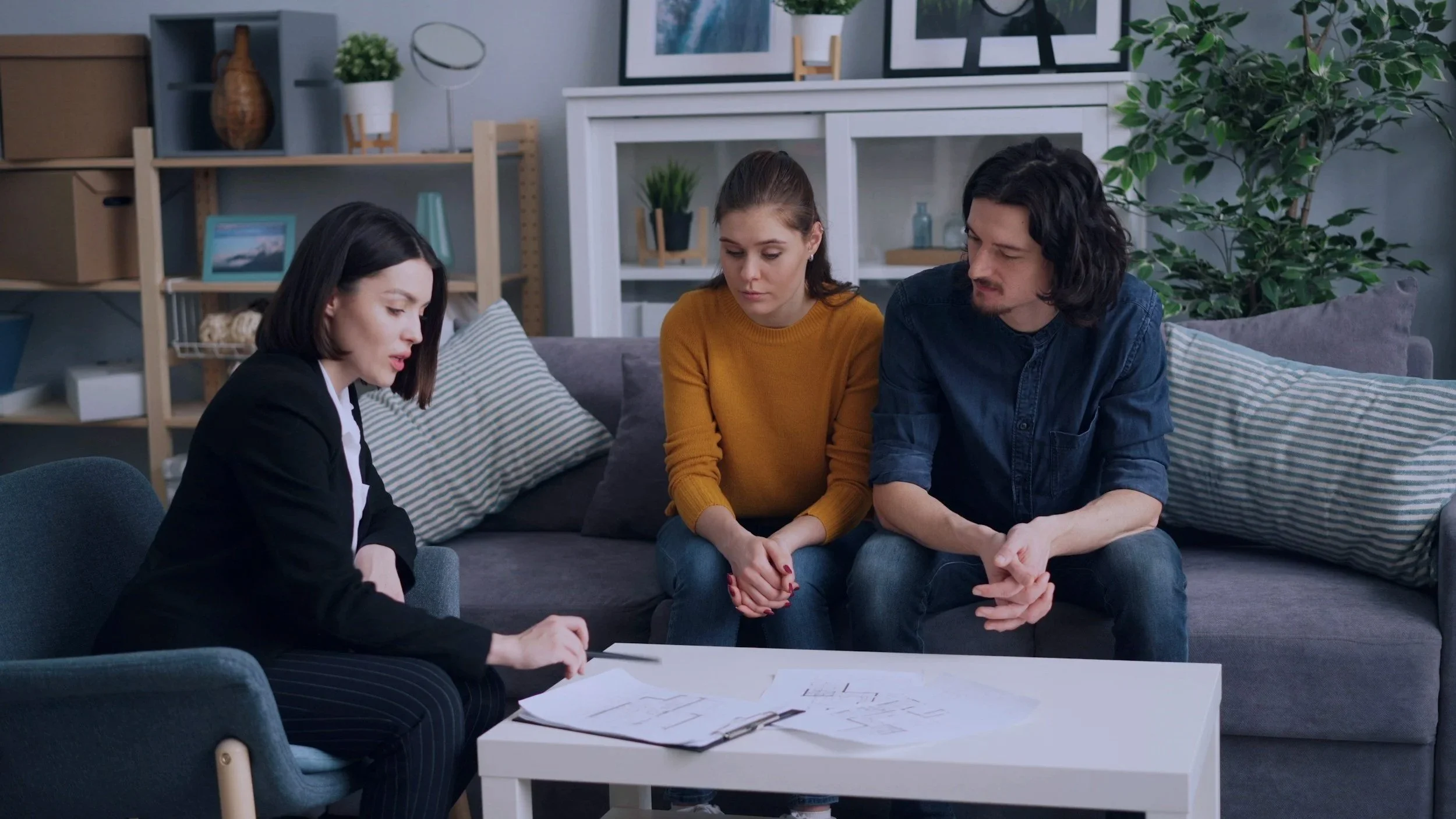Three people sitting on a gray couch and looking at documents on a white table in a living room with shelves, plants, and artwork in the background.