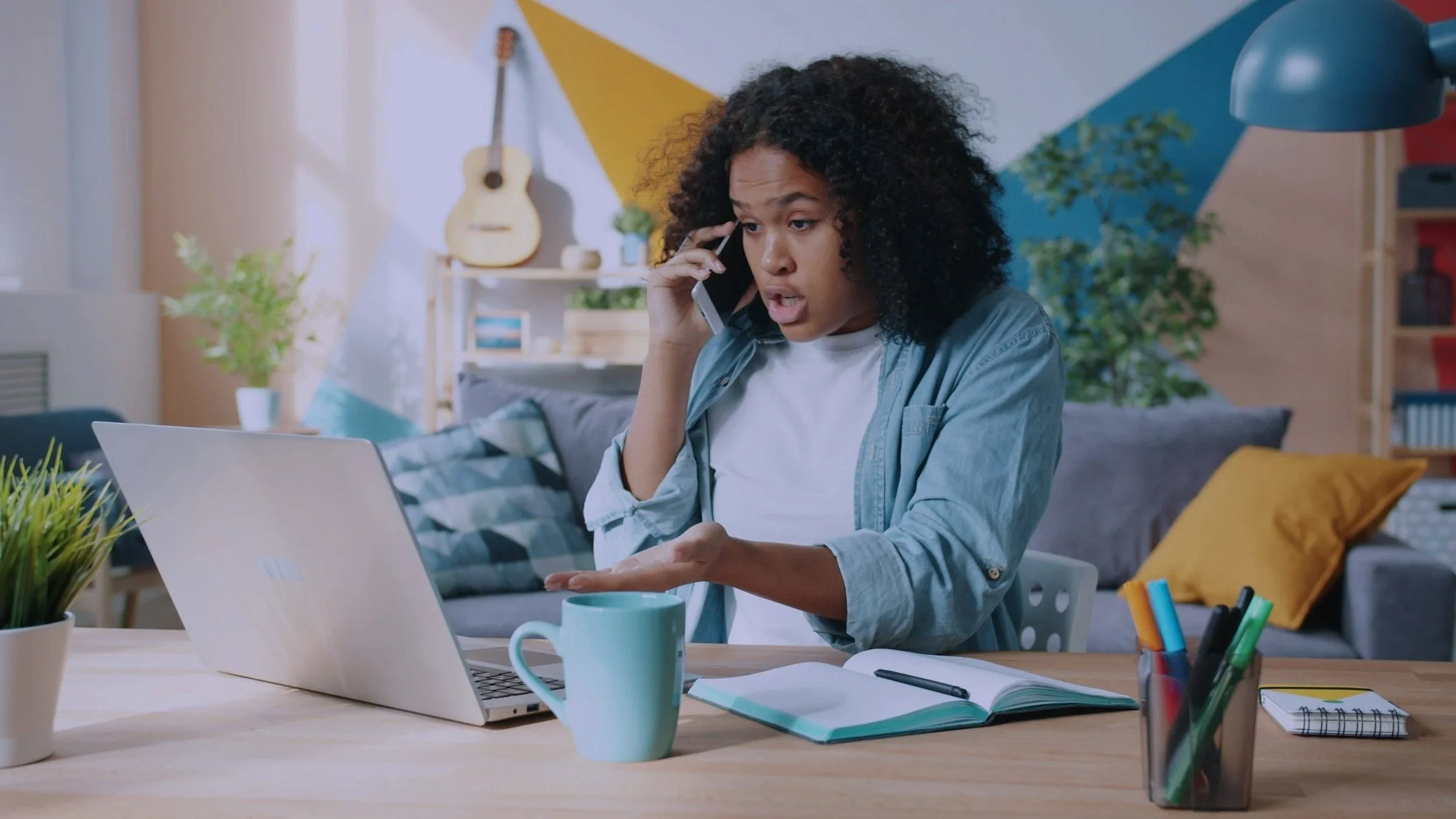 A woman with curly hair talking on her phone while sitting at a desk with an open laptop, a green mug, a notebook, and pens in a colorful and decorated living room.