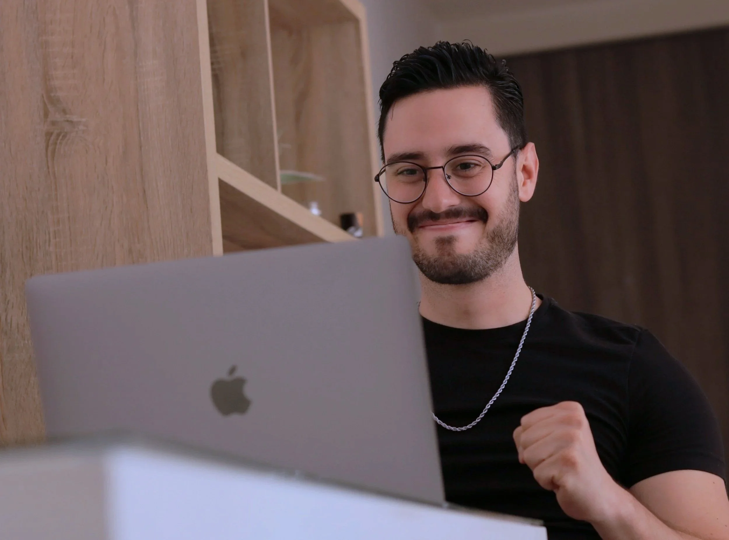 A man with black hair, glasses, and a beard smiling while looking at a computer screen. He is wearing a black shirt and a silver chain necklace. The background features wooden shelves and a dark wall.
