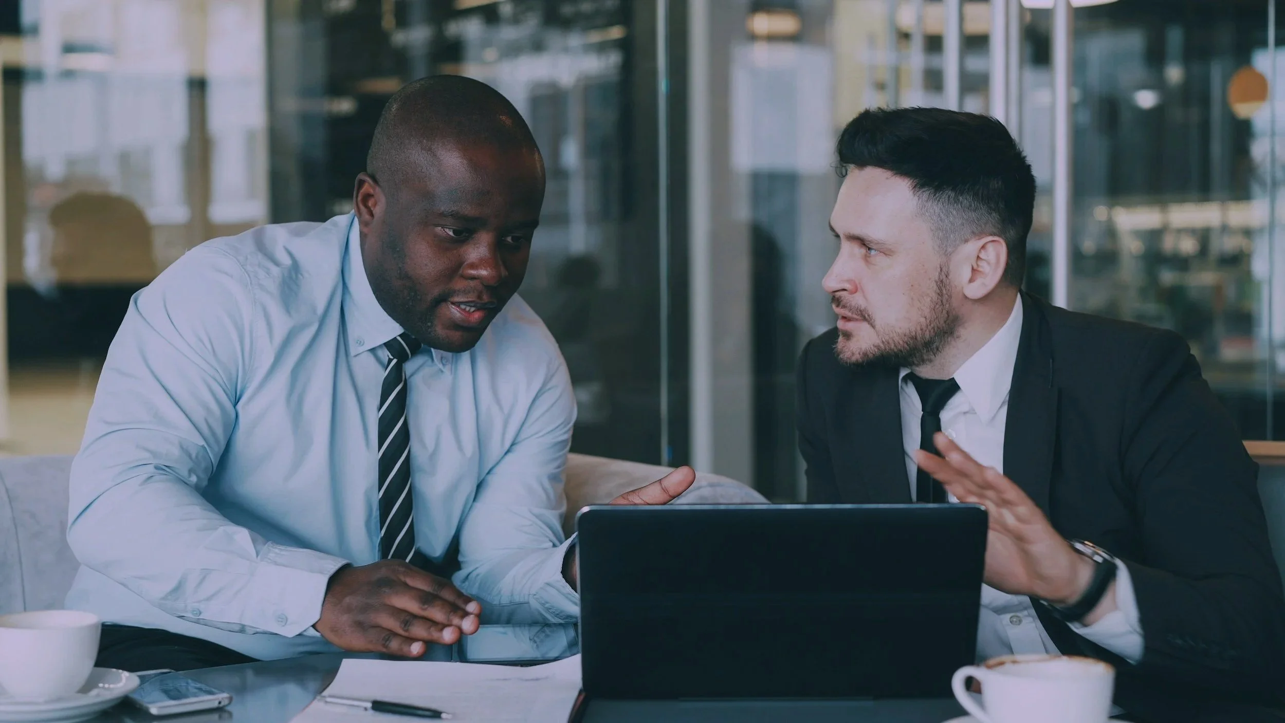 Two businessmen, one Black and one white, in a discussion in a modern office setting. The Black man is wearing a blue dress shirt and a striped tie, while the white man is dressed in a dark suit with a white shirt and black tie. They are seated at a table with a laptop, cups, and documents.