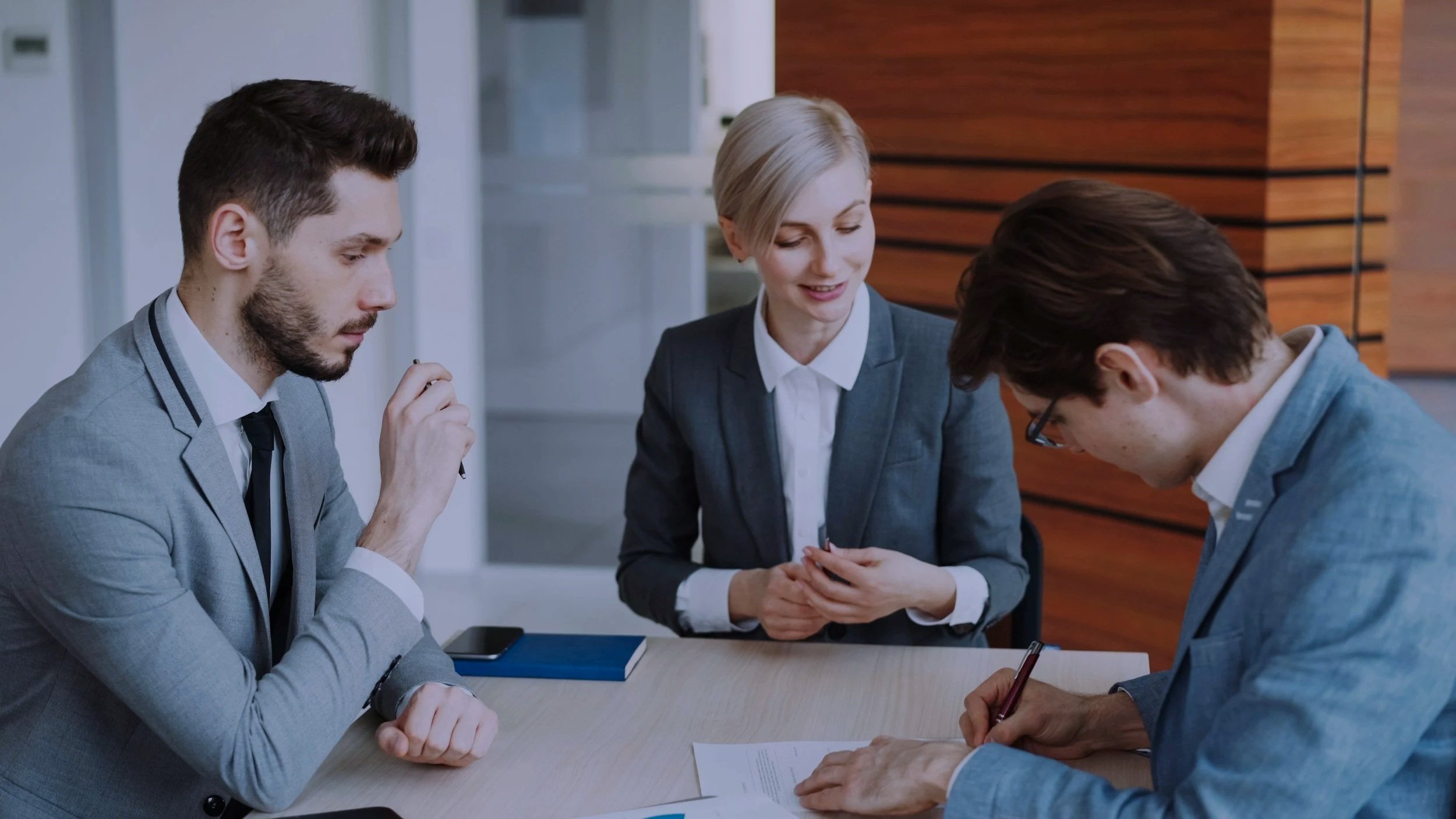 Four professionals in business attire sitting at a table, engaged in a discussion or signing documents, in a modern office setting with wood paneling background.