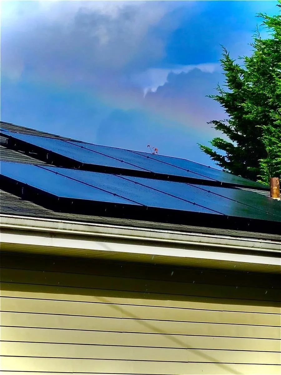A house with black solar panels installed on the roof, a green pine tree on the right, and a cloudy blue sky above.