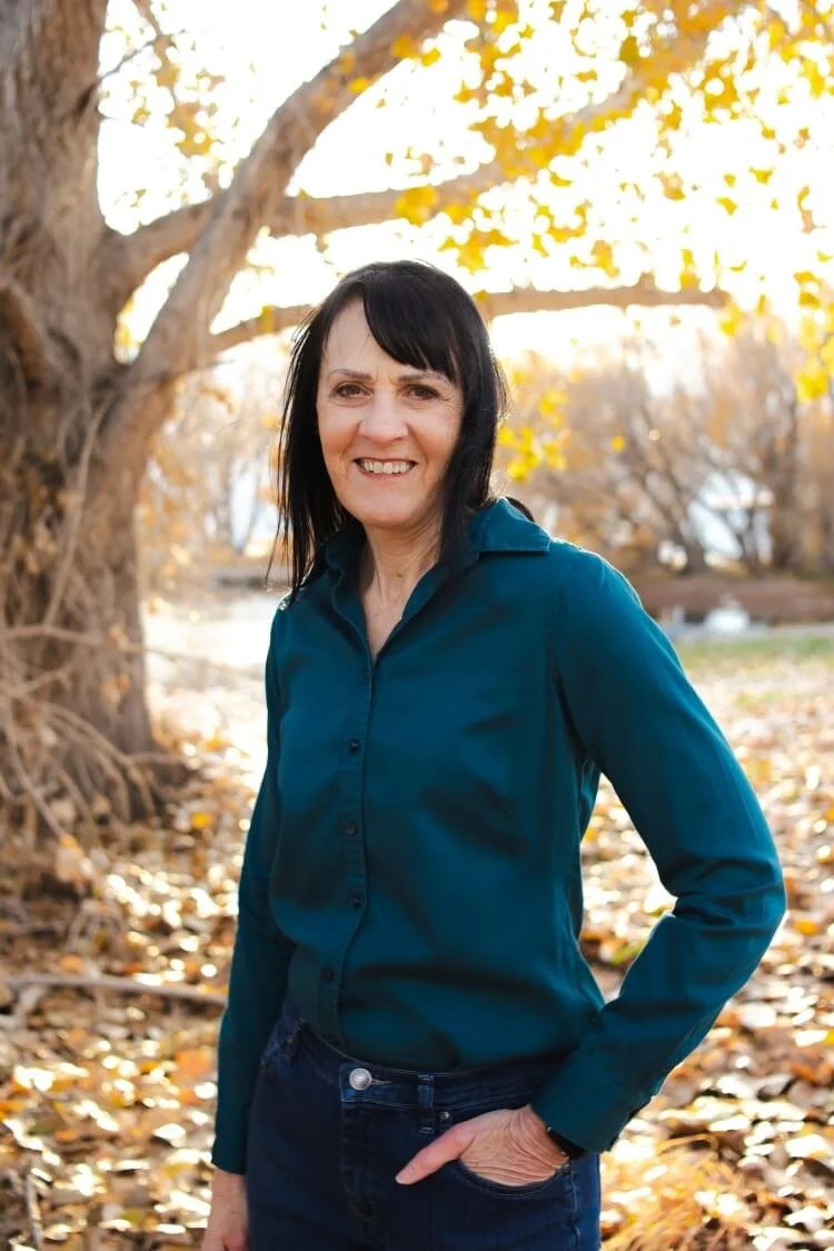 Alison L. Randall wears a teal button-down shirt and stands among fall leaves with a pond in the background. The scene is backlit by golden light.