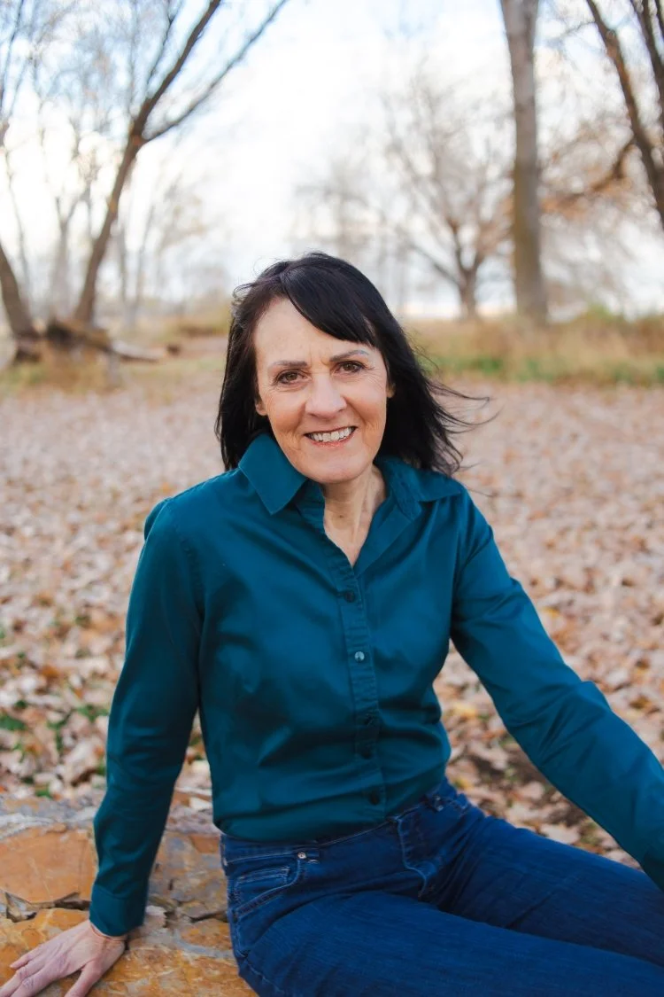 Author Alison L. Randall sits on a rock surrounded by fallen leaves. She wears a teal button-down shirt and a smile.