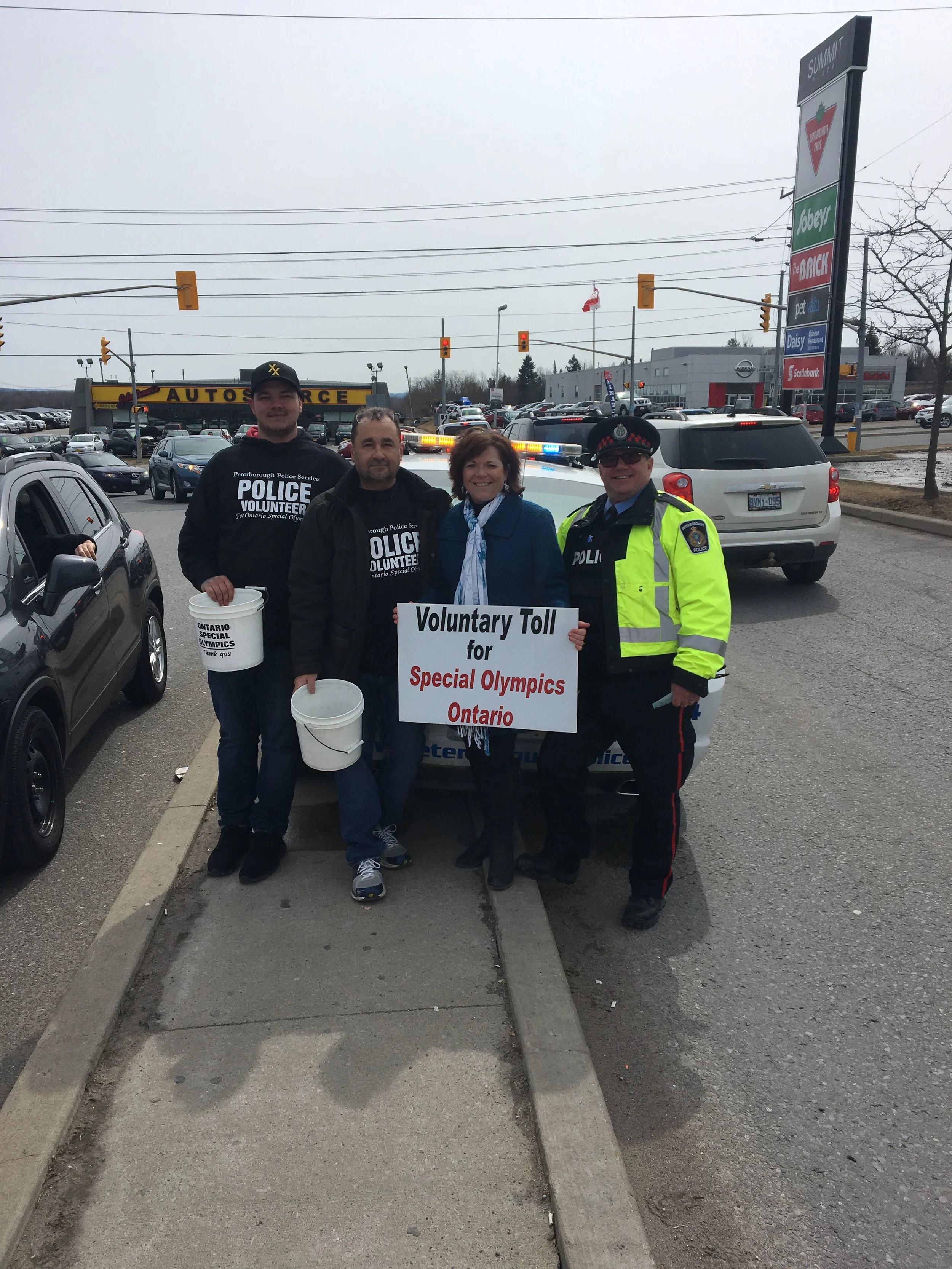 Four people standing on a sidewalk holding signs and buckets at a charity event for Special Olympics Ontario, with cars and a car dealership in the background.