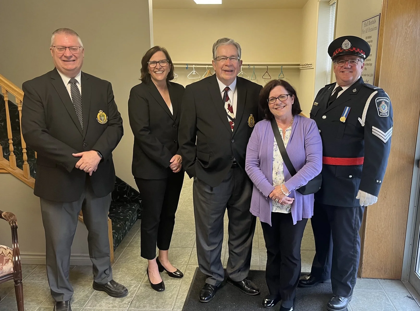 Group of five smiling individuals standing together in an indoor setting. The group includes three men in formal suits, one woman in formal attire, and a police officer in uniform. The background features a yellow wall with a sign and a coat rack wit