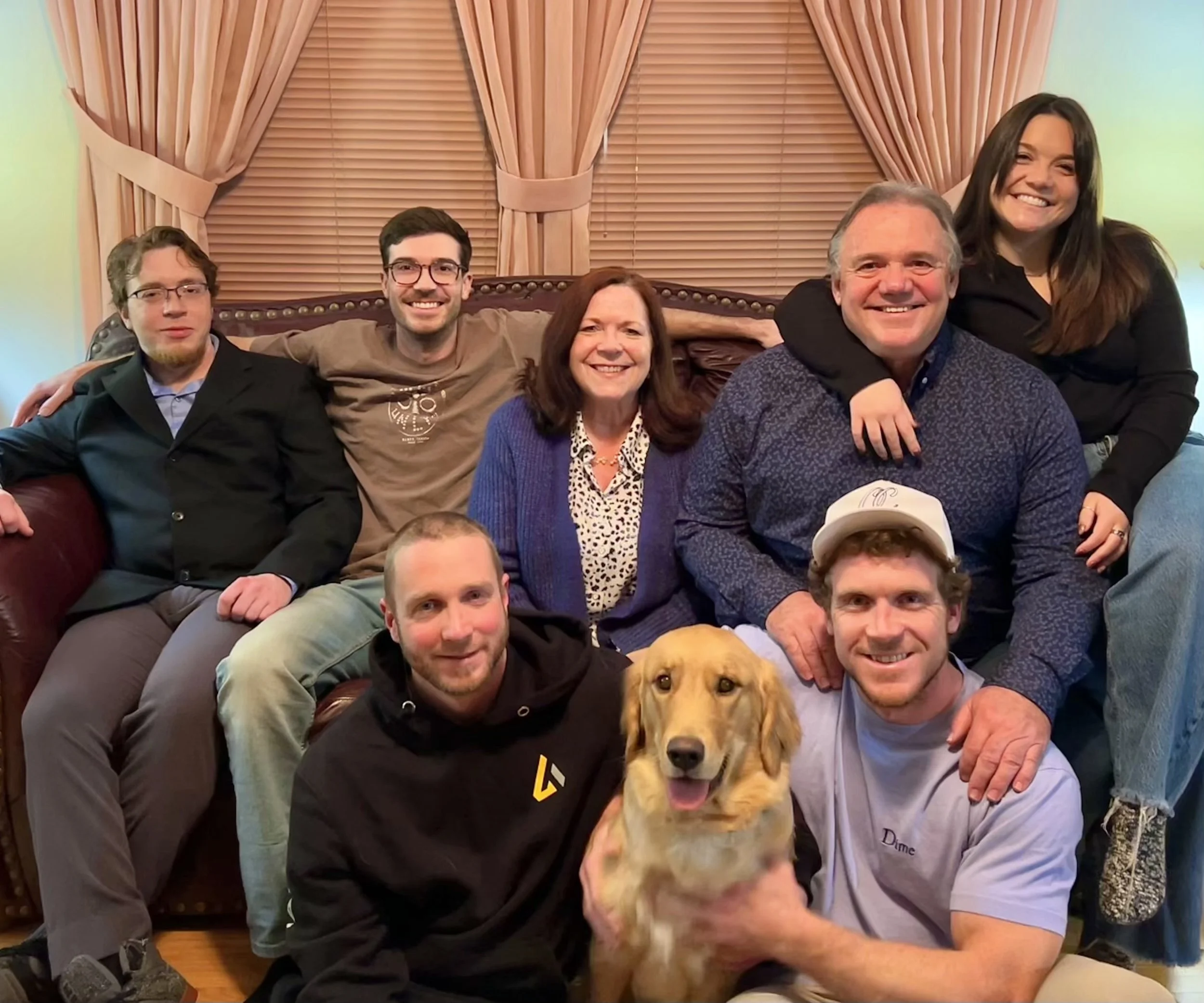 A group of six people sitting on a couch with a dog in front. The background features pink curtains and wooden blinds. The group includes four men and two women, smiling and posing for the photo.