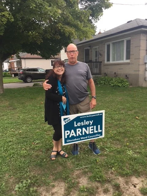 Two people standing outdoors next to a campaign sign for Lesley Parnell, Otonabee Ward Councillor, with a house and trees in the background.