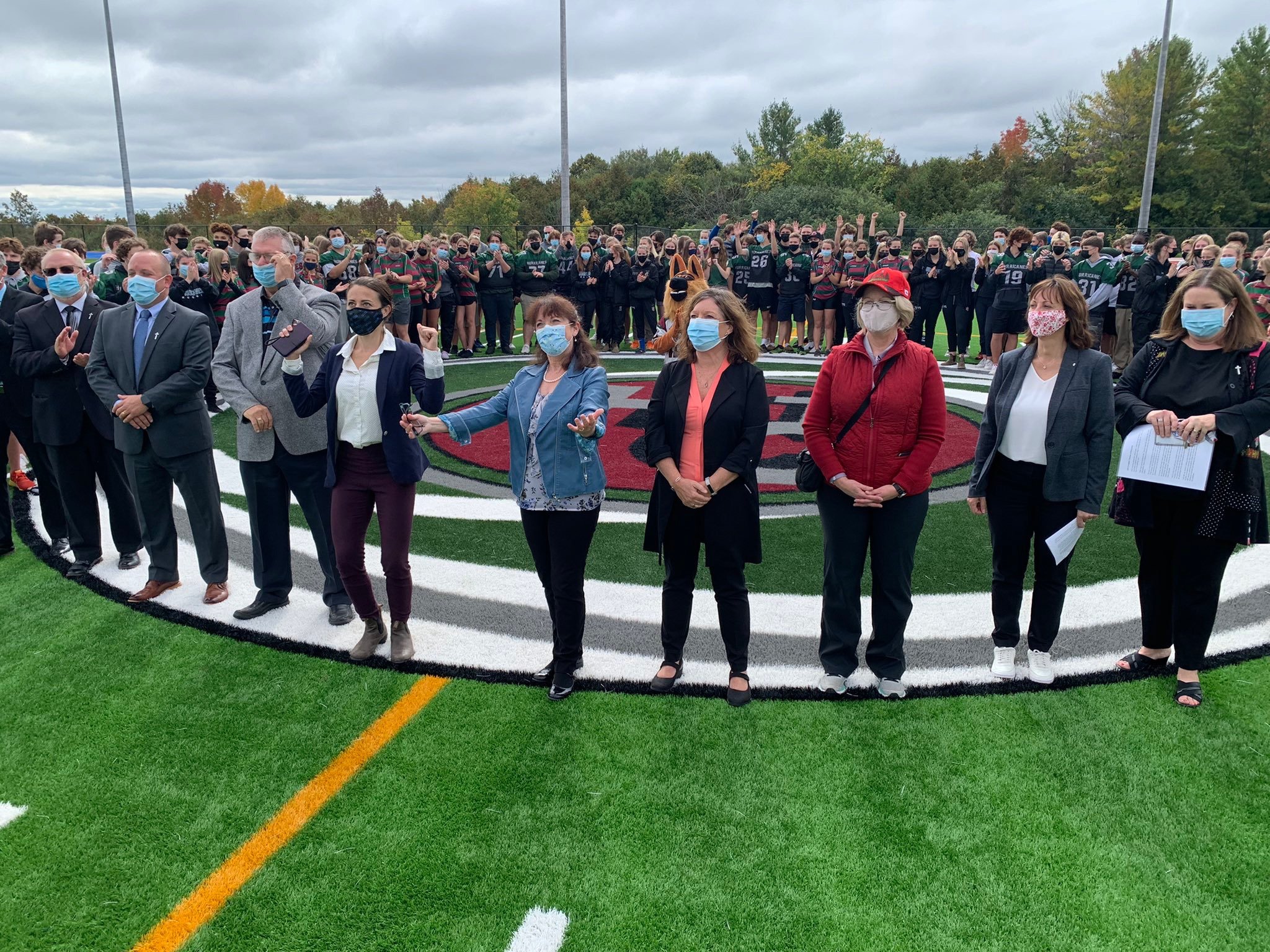A group of people, including students in sports uniforms, standing on a football field with a school logo, wearing masks, and a few adults, possibly teachers or officials, also wearing masks, during an outdoor event on a cloudy day with trees in the 