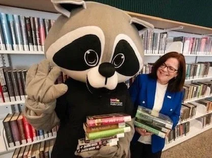 A person dressed in a raccoon mascot costume standing next to a woman in a blue blazer, both holding stacks of books, in front of library bookshelves.