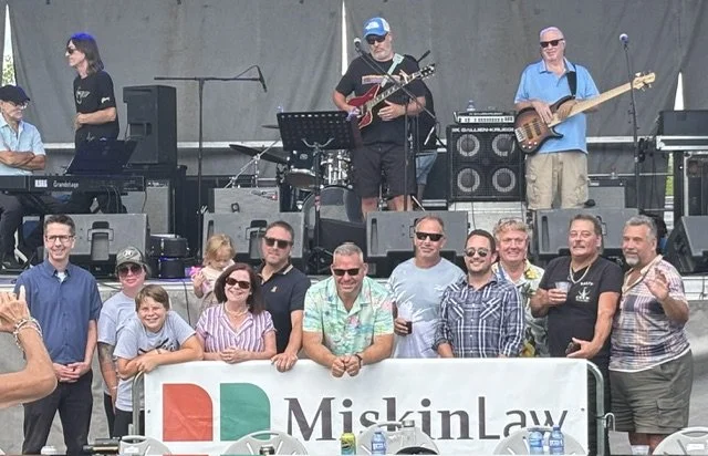 Group of people posing in front of a band performing live music, with a banner reading 'Mishin Law' in front.