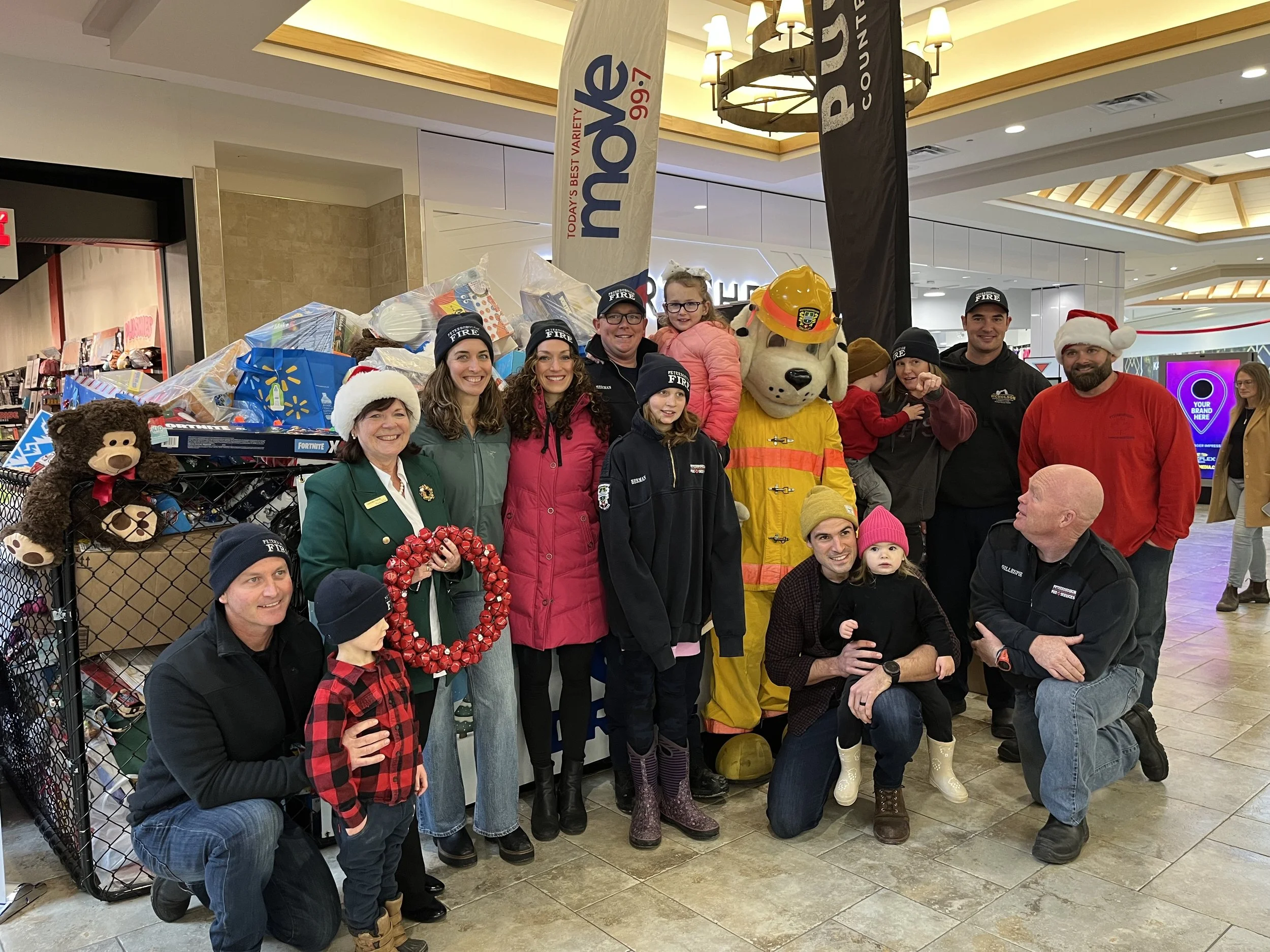 A group of people including adults, children, and a person in a dog mascot costume, gathered in a shopping mall, some wearing Christmas hats and holding holiday decorations, posing for a photo during a Christmas event.