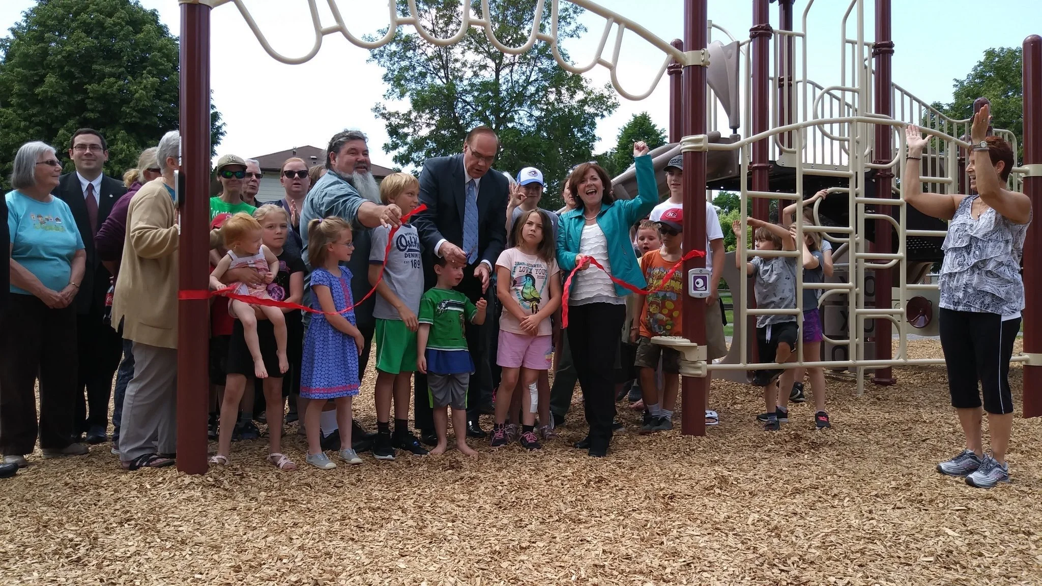 Group of children and adults at a ribbon-cutting ceremony for a new playground, some children hold the ribbon, and a woman is about to cut it while others watch and smile.