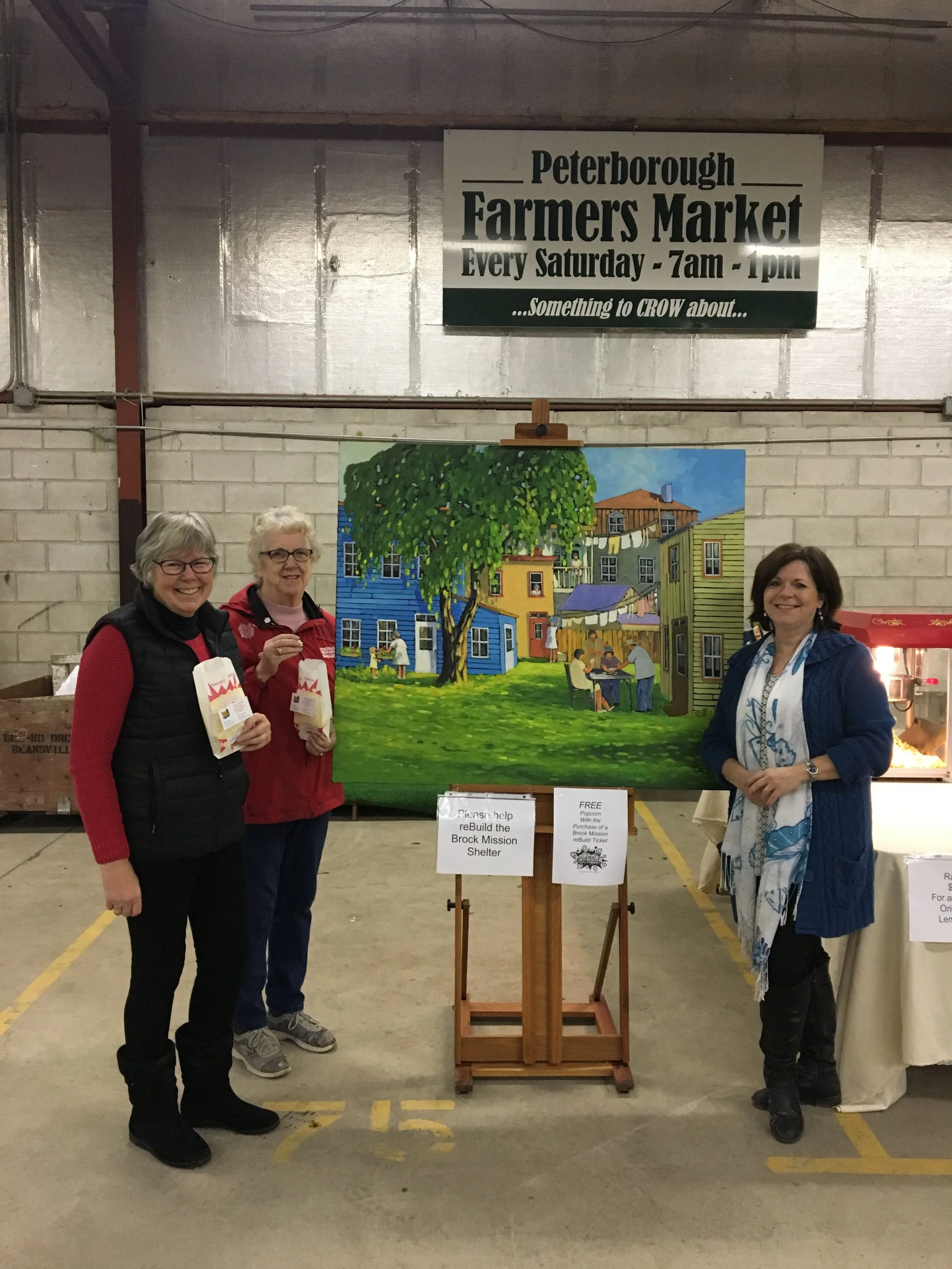 Three women standing in front of a colorful painting of a neighborhood scene with houses, trees, and people, inside a building with a sign advertising a farmer's market. The women are smiling, holding popcorn, and wearing casual clothes.