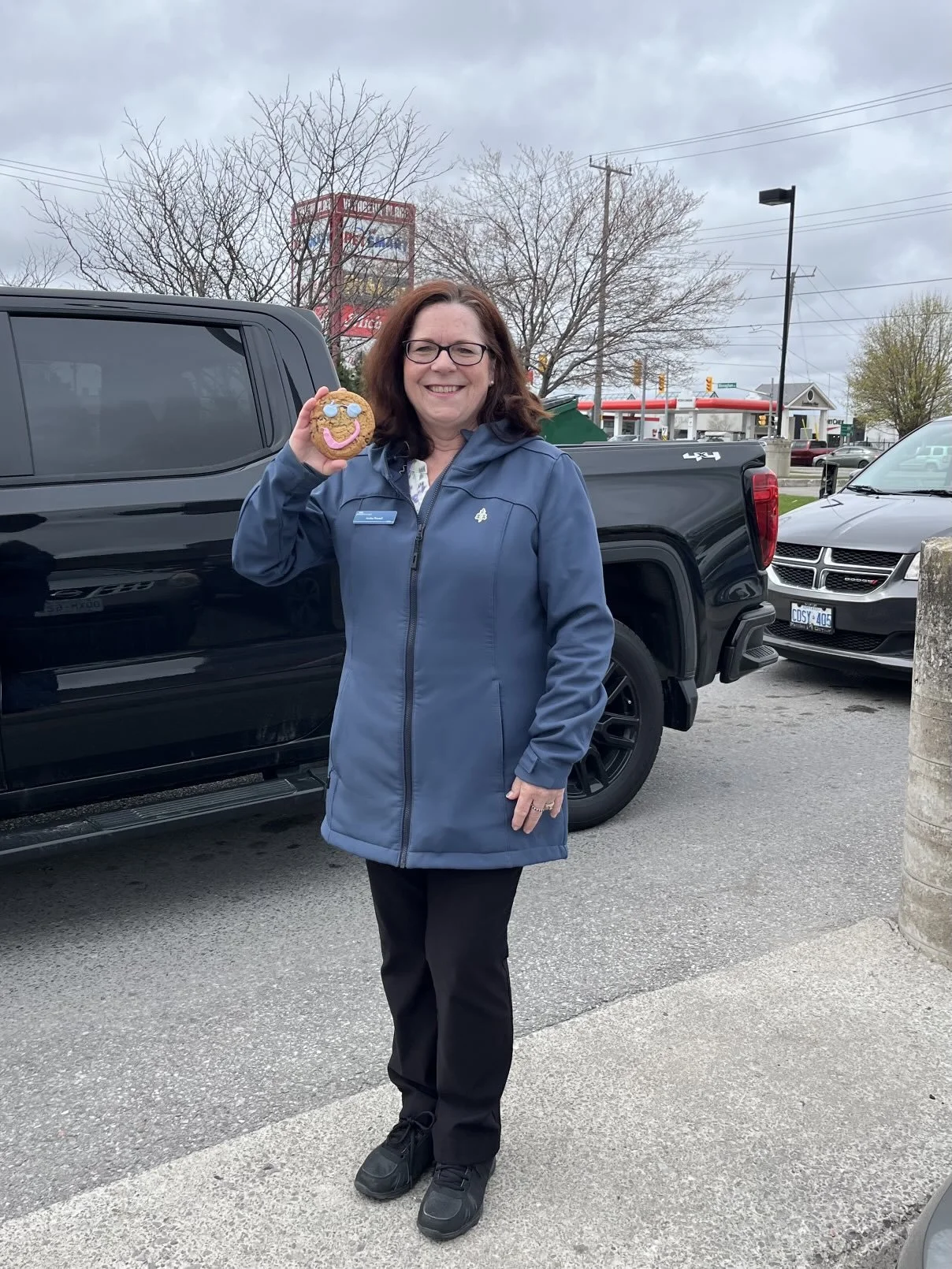 A smiling woman wearing glasses and a blue jacket standing outside in a parking lot, holding a cookie with a smiley face drawn on it.