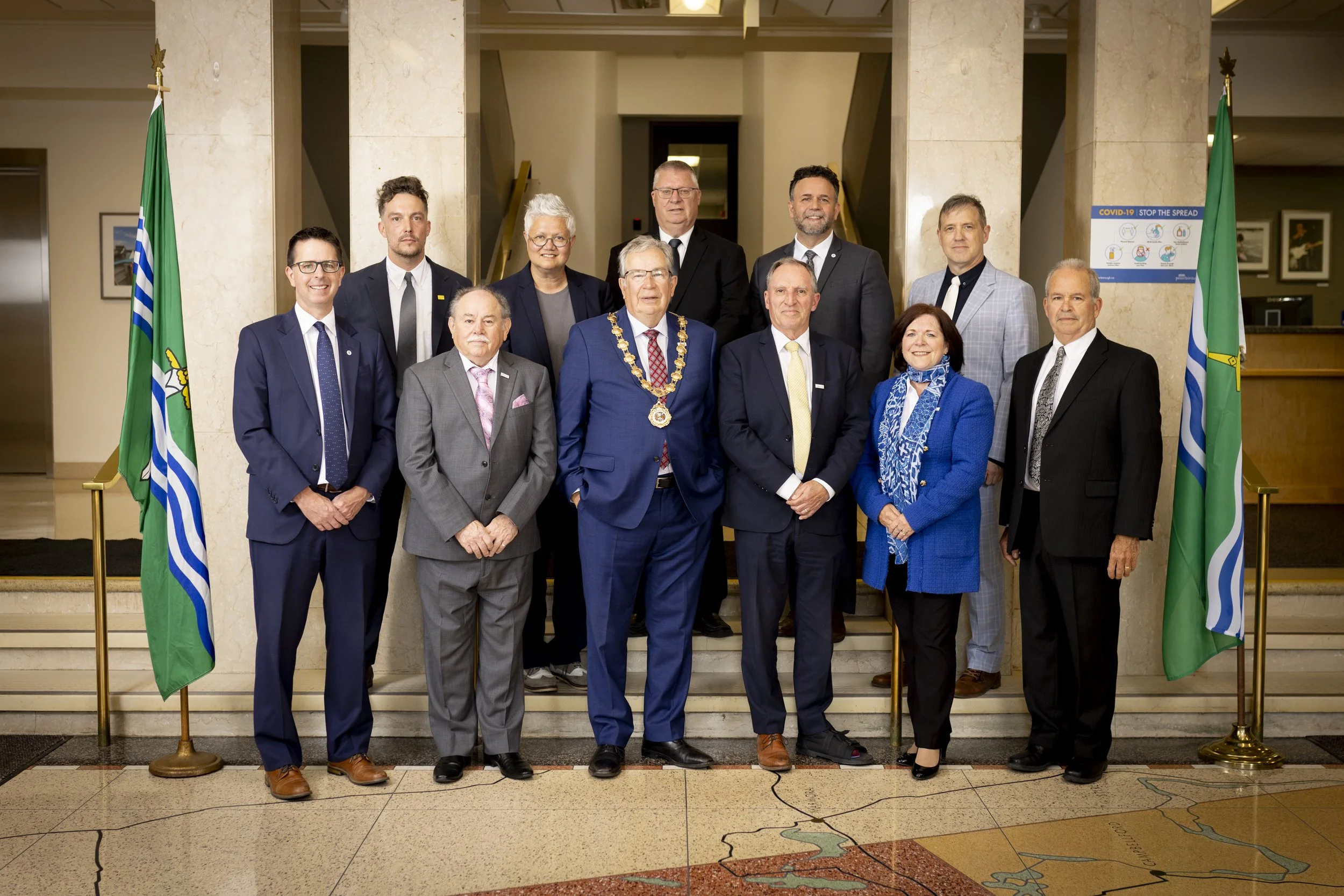 A group of fourteen people dressed in formal attire standing on a staircase for a formal photograph. The central figure wears a blue suit and a mayoral chain. Two flags flank the group on either side, and there is a COVID-19 informational poster on the wall behind them.