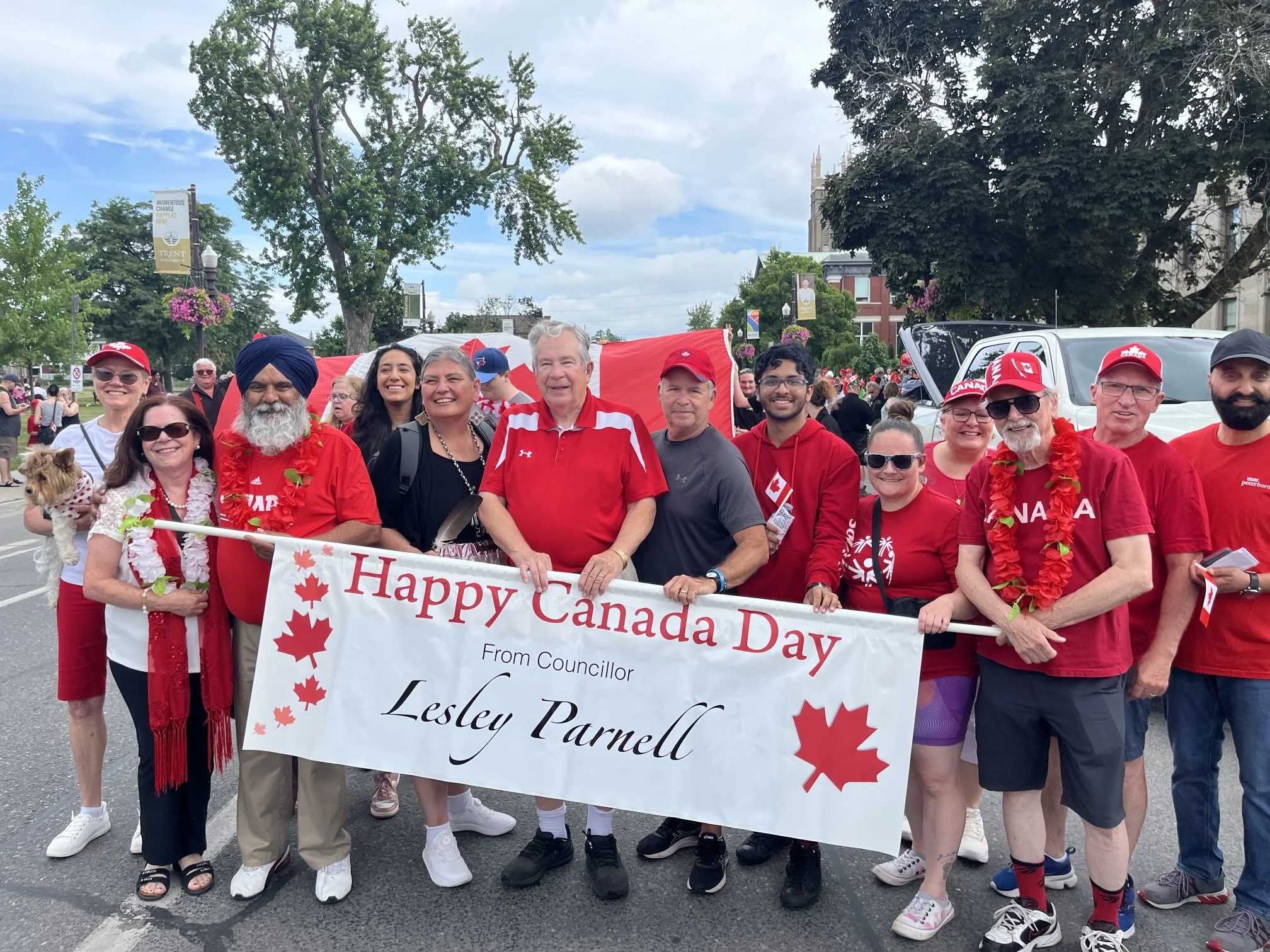 Group of people participating in a Canada Day parade, holding a banner that says 'Happy Canada Day' with maple leaf symbols. They are dressed in red and white, some wearing hats, leis, and sunglasses. Some people are smiling, and a small dog is visib