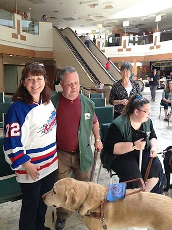 Four people and a guide dog in a shopping mall. Two women are smiling at the camera, one man stands next to them, and a woman seated with a cane is looking away. The guide dog is lying down with a blue vest.