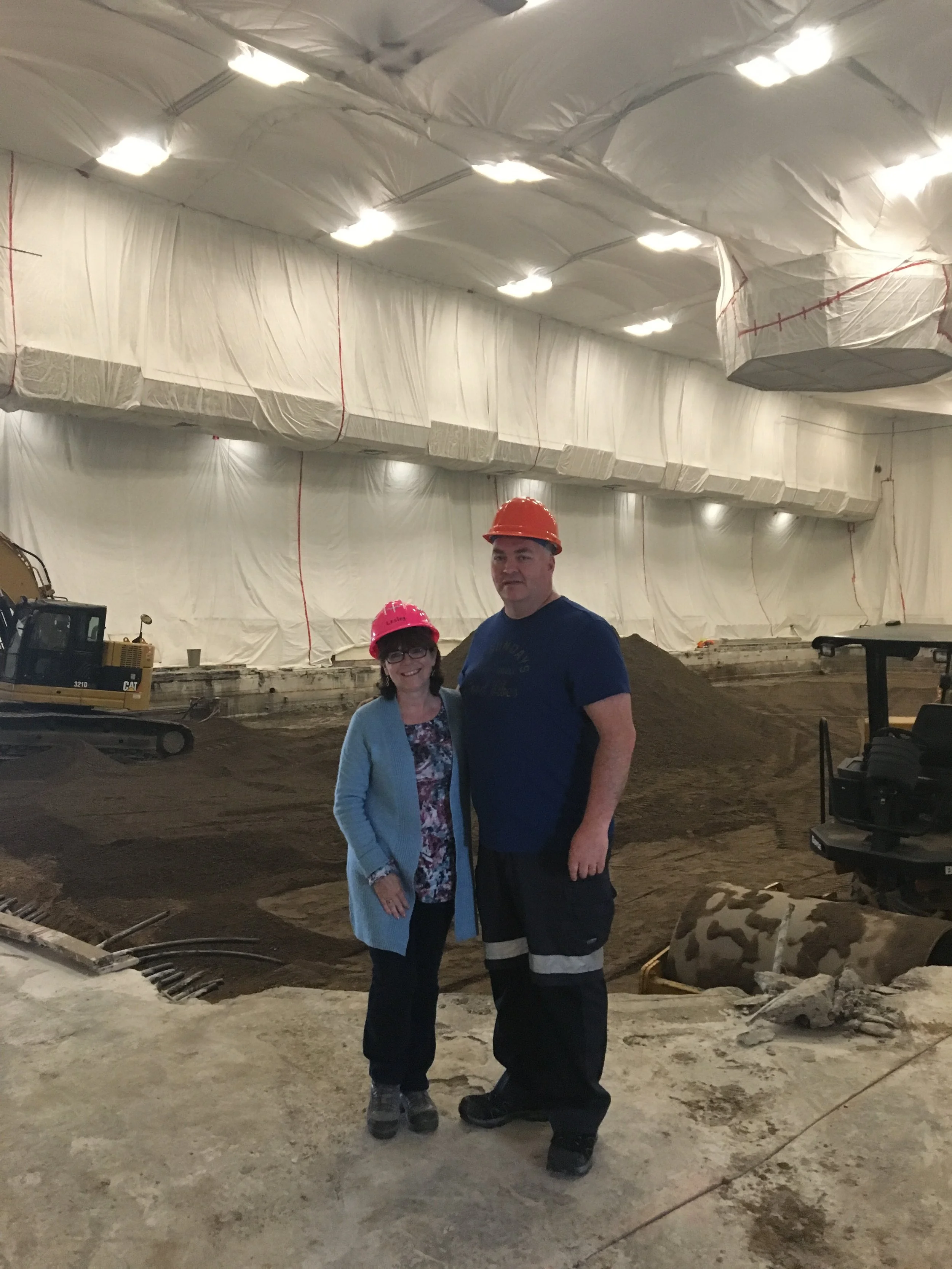 Two people wearing safety helmets standing inside a construction site with equipment and materials, under a partially covered ceiling.
