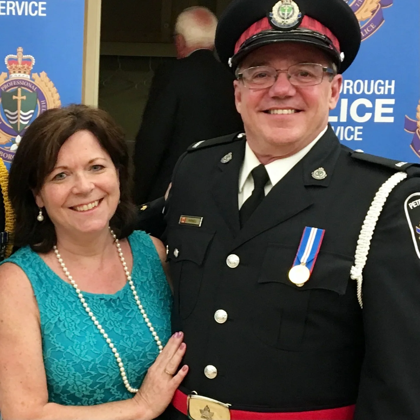 A woman in a teal dress with pearl jewelry standing next to a smiling police officer in uniform at a formal event.