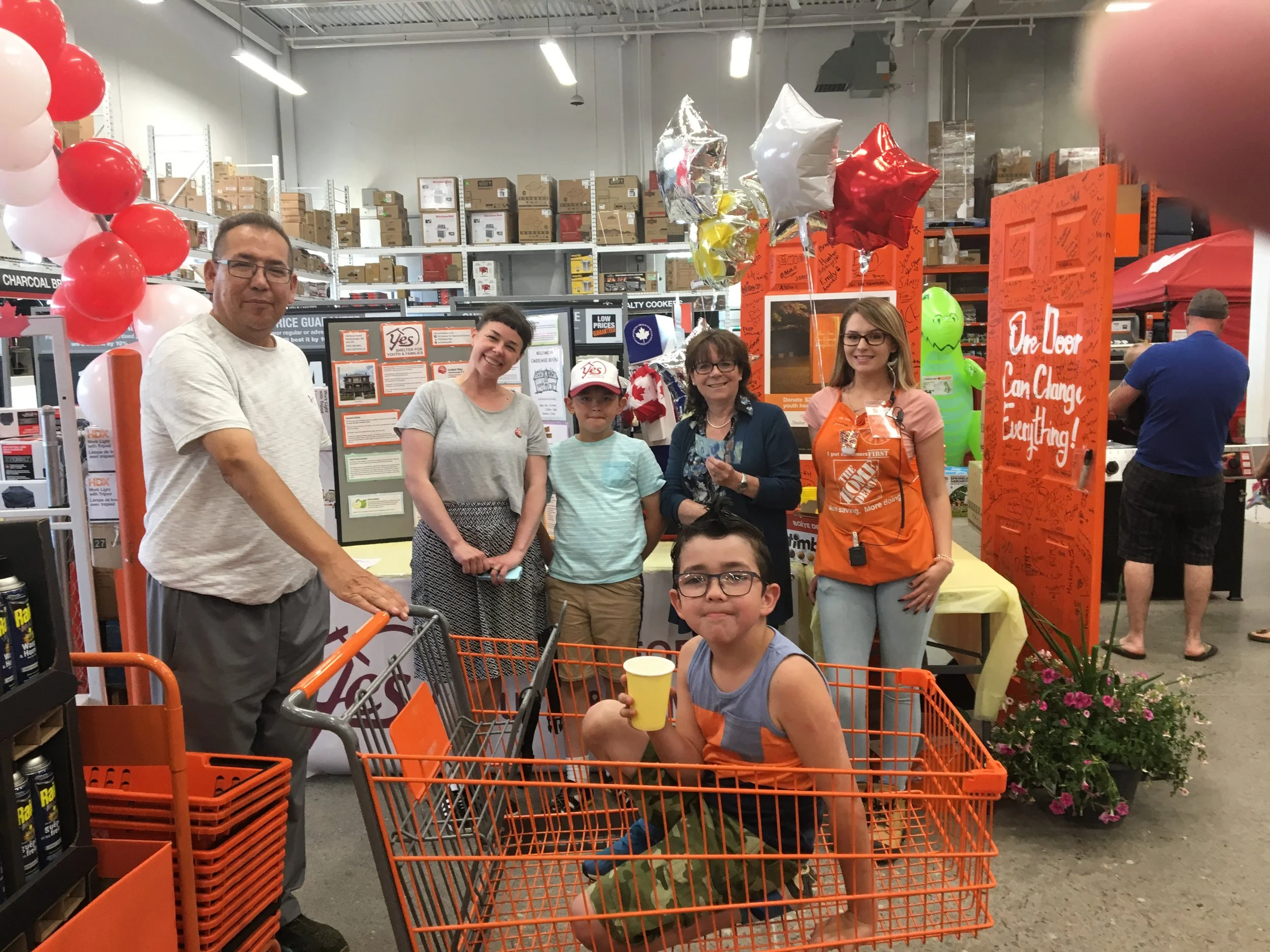 A group of five people, including children and adults, pose for a photo inside a warehouse-like store next to shopping carts. They are smiling and standing in front of a display with orange signage and balloons, surrounded by shelves stocked with box
