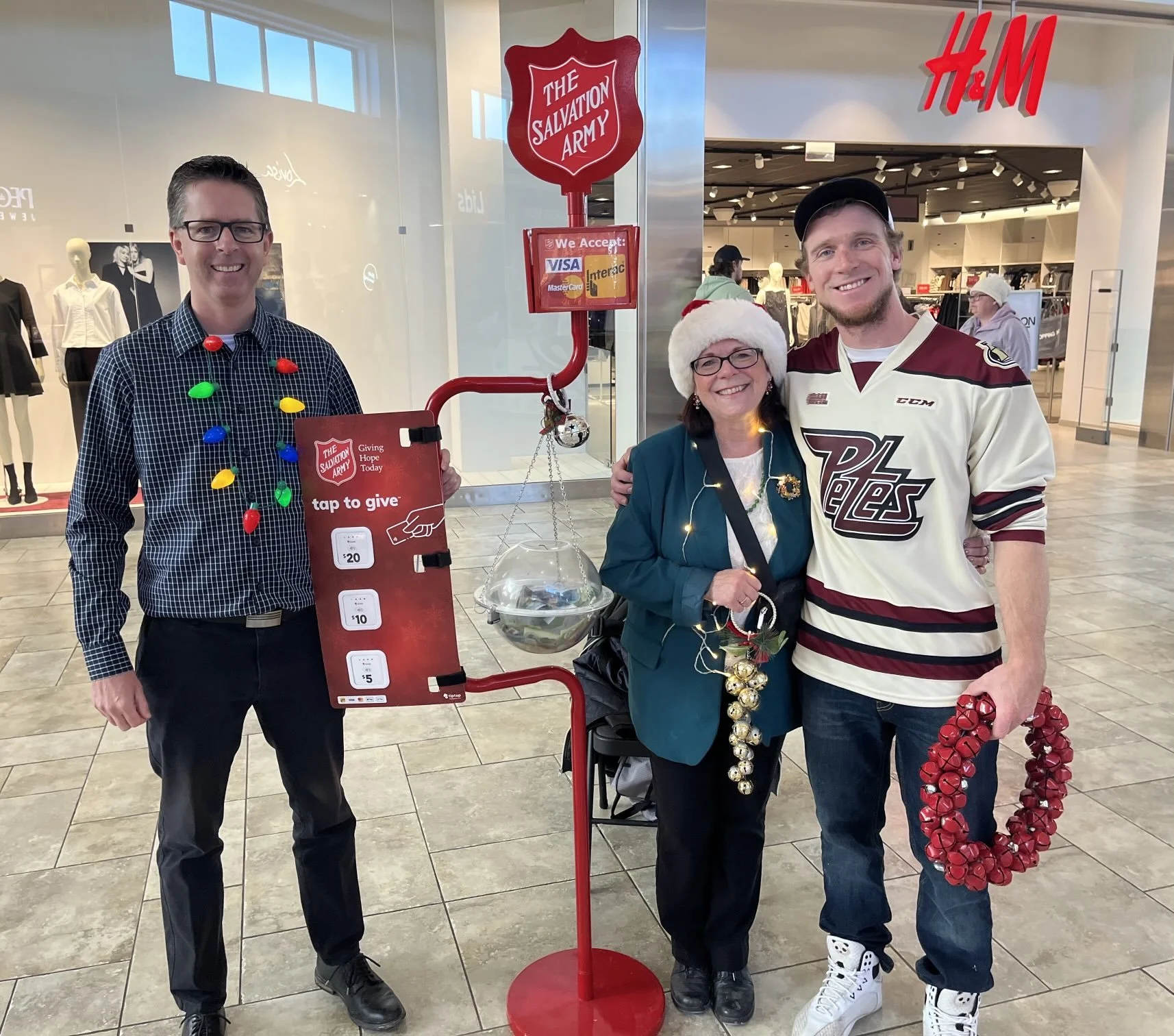 Three people standing together in a shopping mall decorated for Christmas, with a Salvation Army donation stand behind them. One man on the left is wearing glasses, a checkered shirt, and holiday lights necklace. In the middle, a woman wearing glasse
