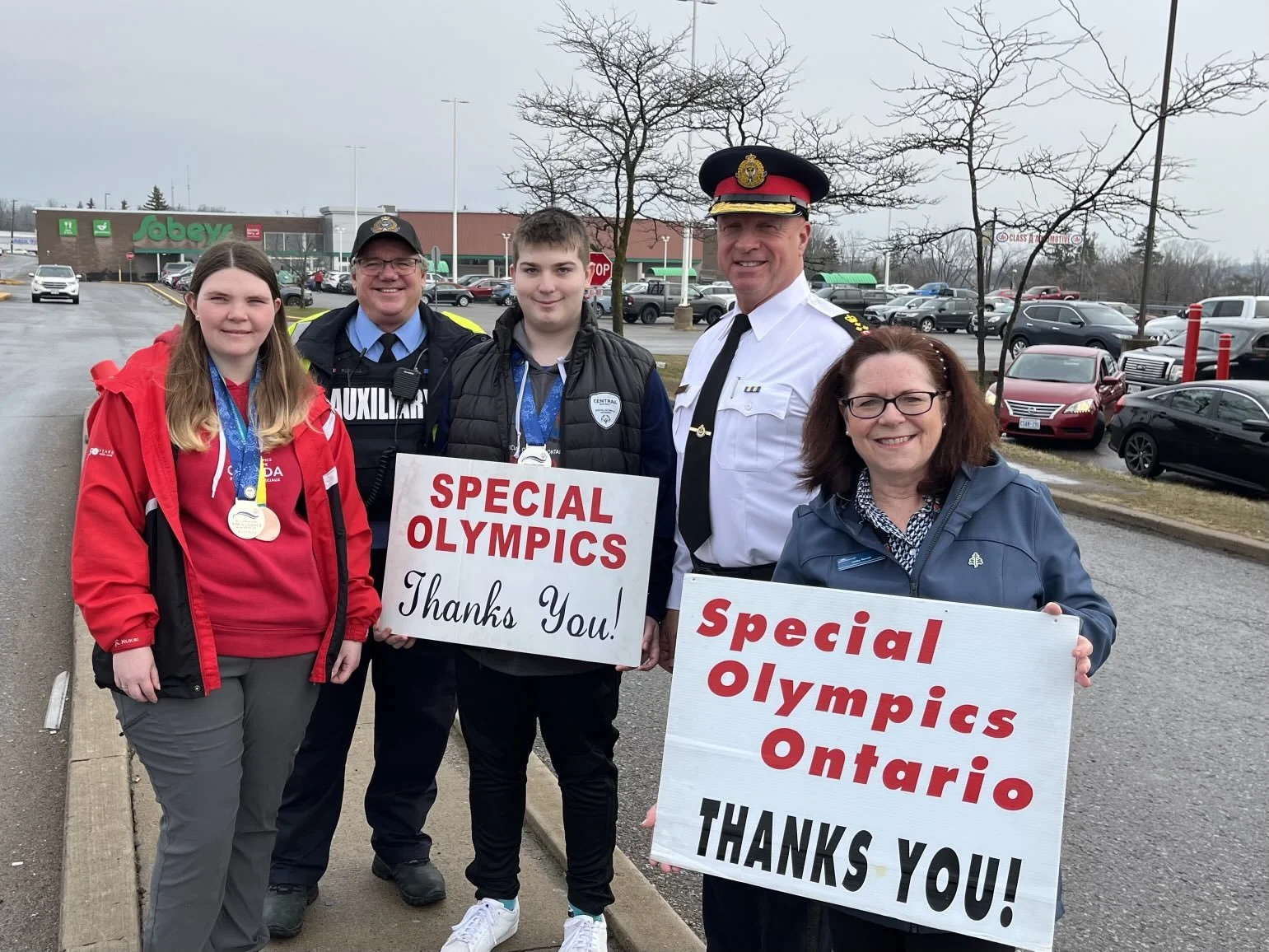 A group of five people standing outdoors in a parking lot, holding signs that thank Special Olympics Ontario and Ontario organizers, with medals around some of their necks.
