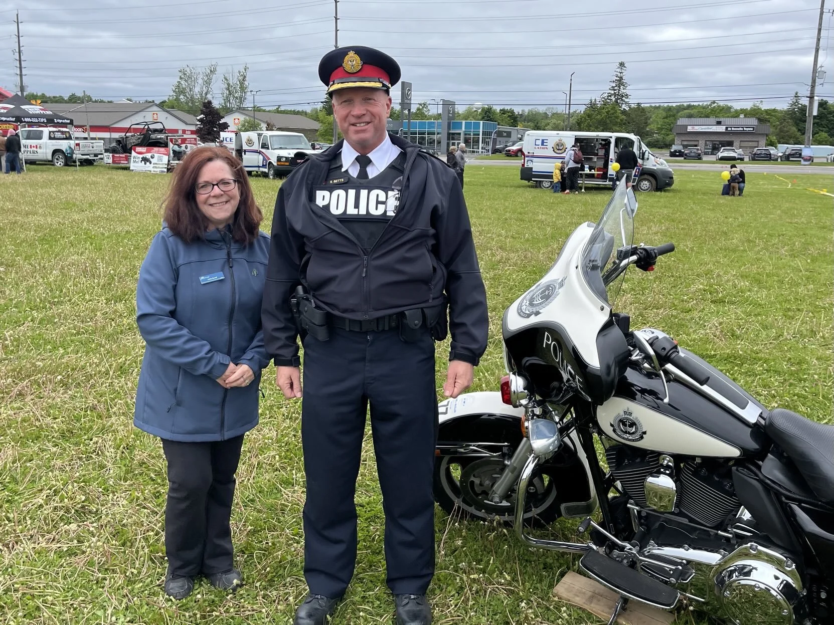 A woman and a police officer standing outdoors on a grassy field, with police vehicles and an event setup in the background, and a police motorcycle parked beside them.