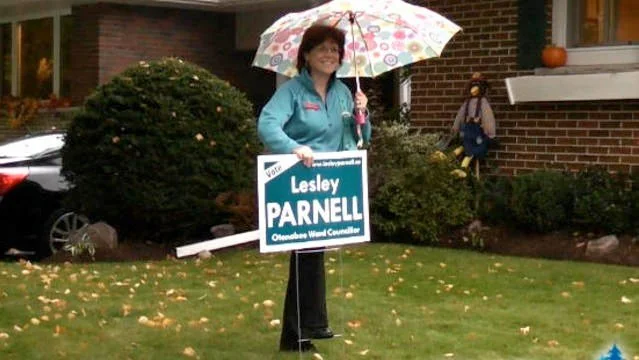A woman holding an umbrella and campaign sign standing on a lawn with fallen leaves in front of a brick house.