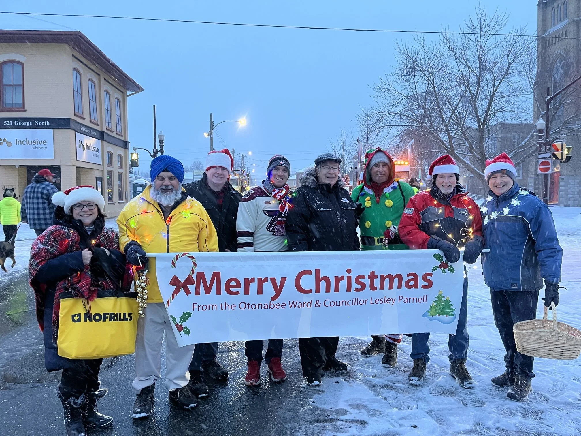 People participating in a Christmas parade on a snowy street, wearing festive hats and costumes, holding a banner that says "Merry Christmas" with a Christmas tree graphic, in a town with buildings and streetlights.
