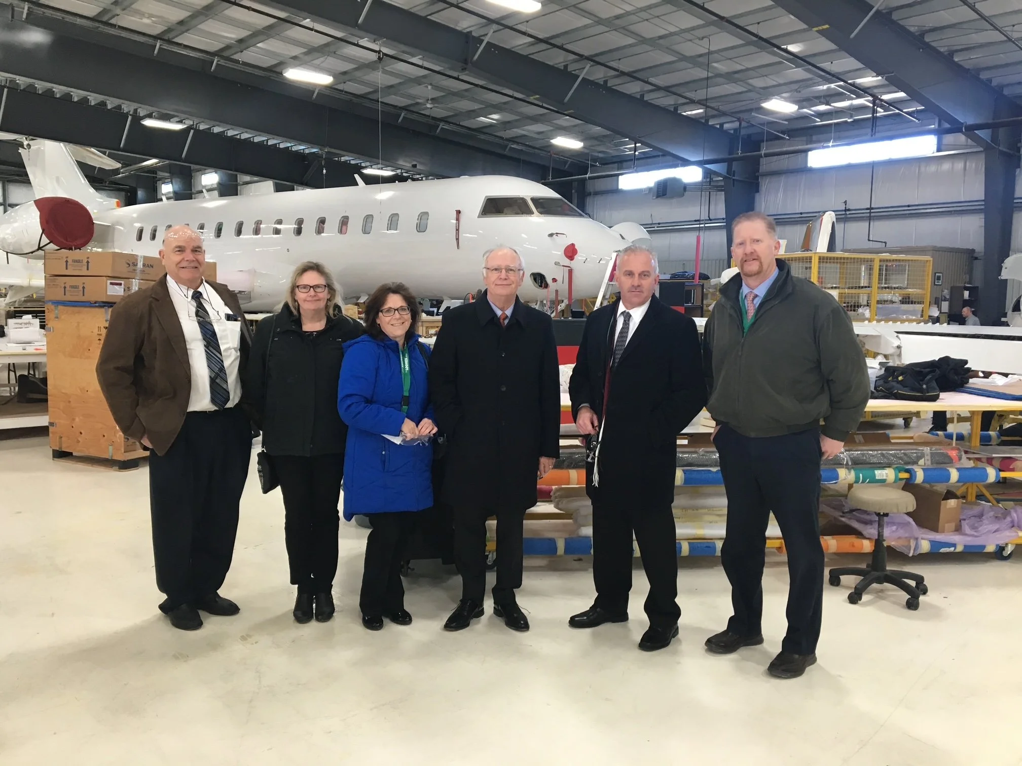 A group of six people standing in front of an airplane inside an industrial hangar or warehouse. The airplane is white and appears to be under construction or maintenance, with tools and equipment around it.