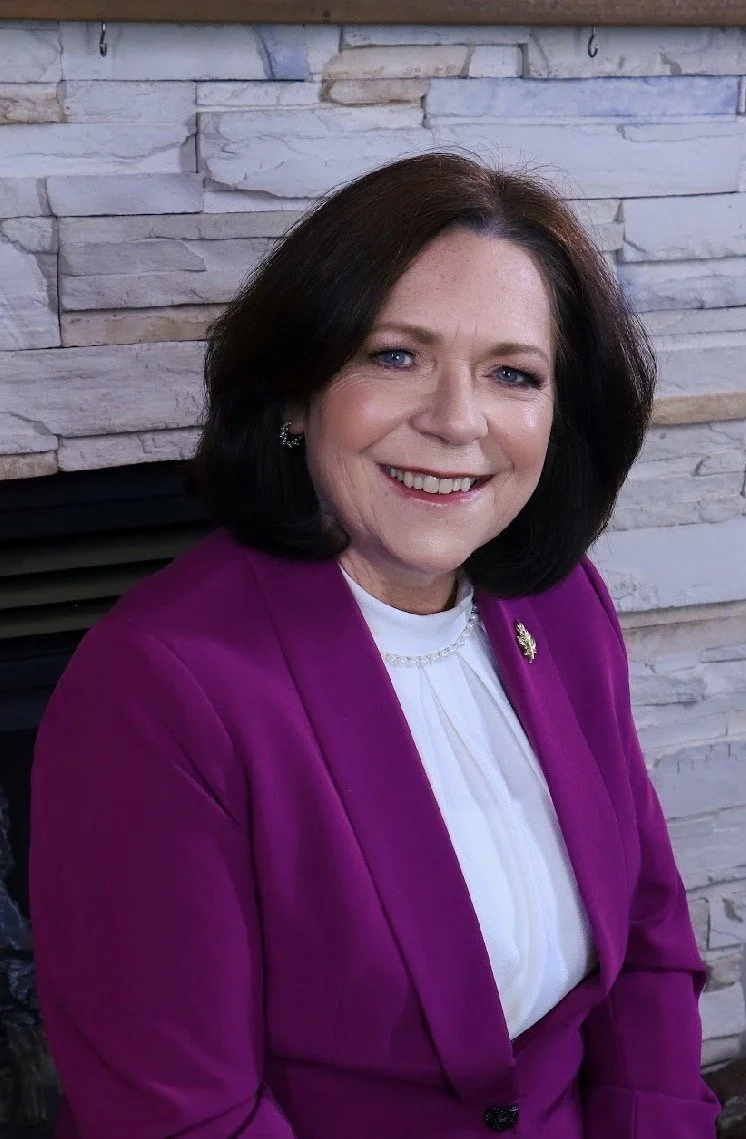 A smiling woman with shoulder-length dark hair wearing a purple blazer and a white blouse, sitting in front of a stone wall.