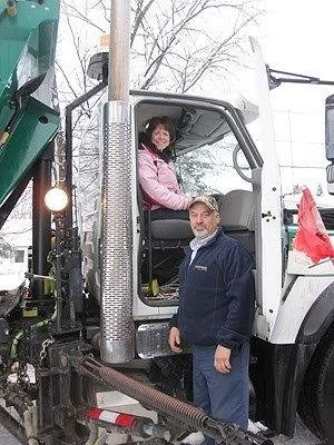 A woman sitting in the driver's seat of a large white truck, smiling, with a man standing outside next to the door, in an outdoor setting with snow and leafless trees.