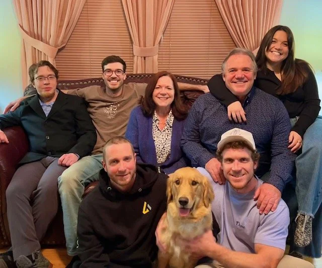 A family of seven people and a golden retriever dog sitting on a living room sofa and floor, smiling and posing for a group photo.