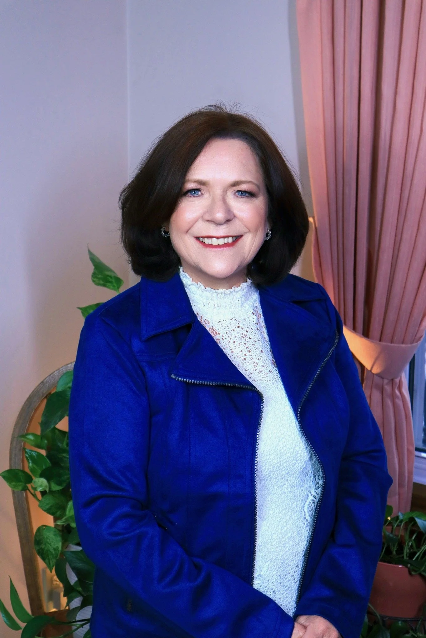 Portrait of a woman with shoulder-length dark brown hair, wearing a white lace top and a blue jacket, smiling in front of a pink curtain and green plants.