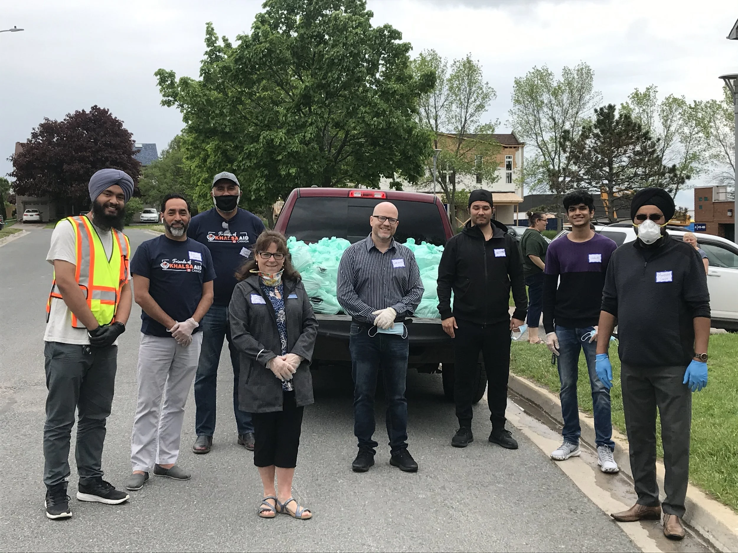 A group of eight people standing outdoors in front of a city background. They are dressed casually, with some wearing face masks and gloves. Behind them, a vehicle is filled with light blue bags. The scene suggests a community service or volunteer ev