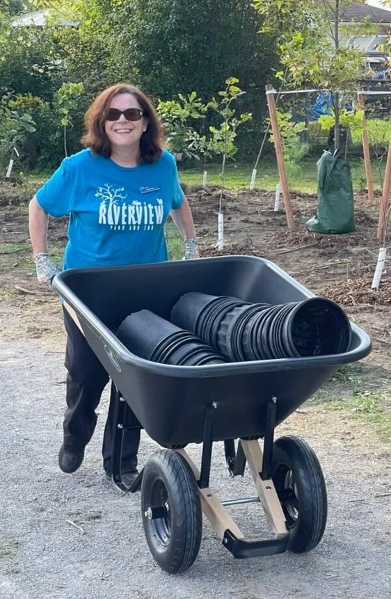 A woman with shoulder-length brown hair, wearing sunglasses, a blue 'Riverview' T-shirt, and gloves, smiling while holding a black wheelbarrow filled with black plant pots outdoors in a garden or park.