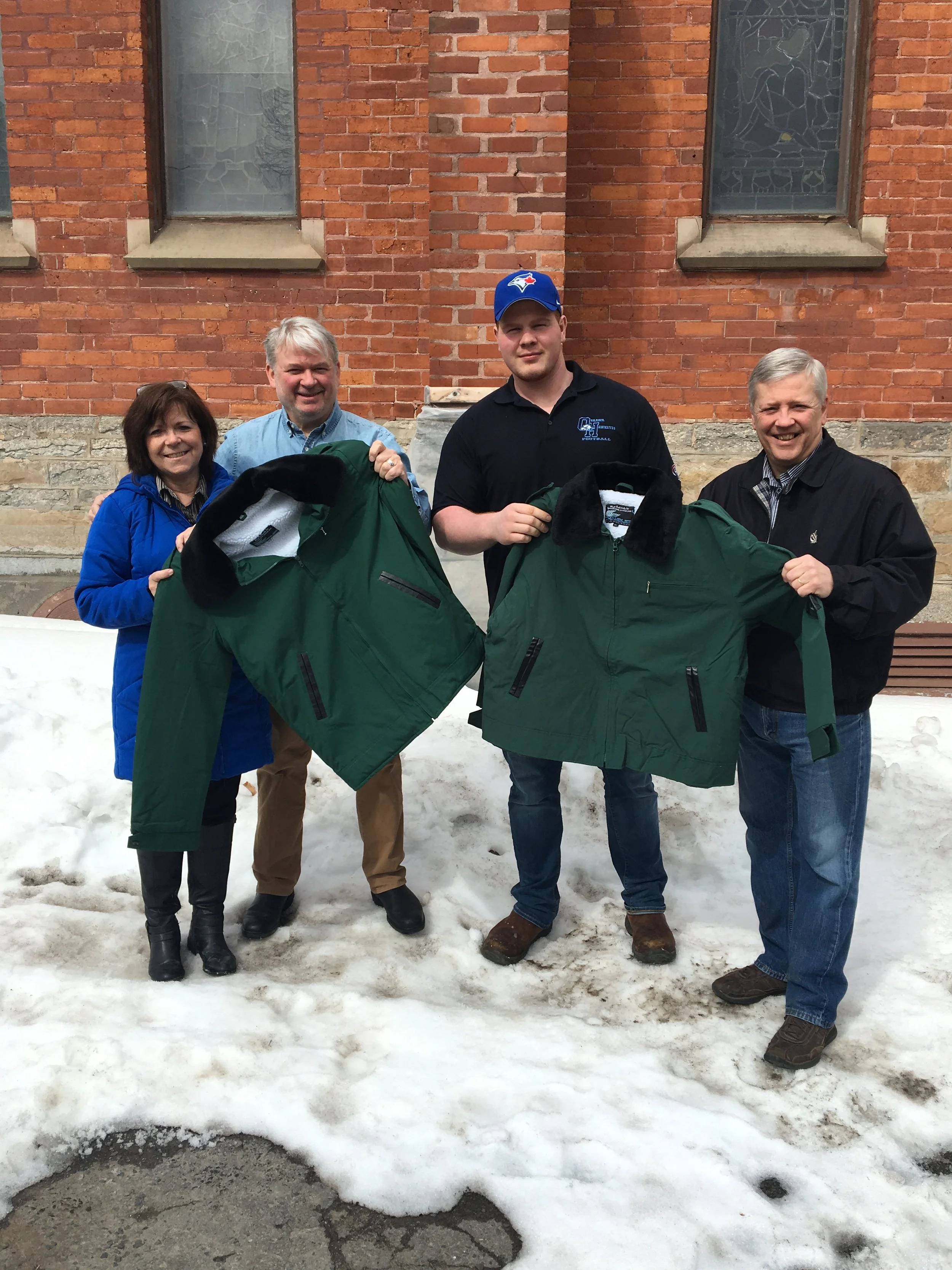 Four adults standing outside on snow in front of a brick building, holding green jackets, smiling, with two windows behind them.